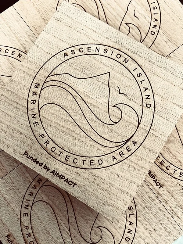 Wooden plaque with a circular logo of a rising wave and the words 'Ascension Island Marine Protected Area' engraved around it, and 'Funded by AIMPACT' beneath.