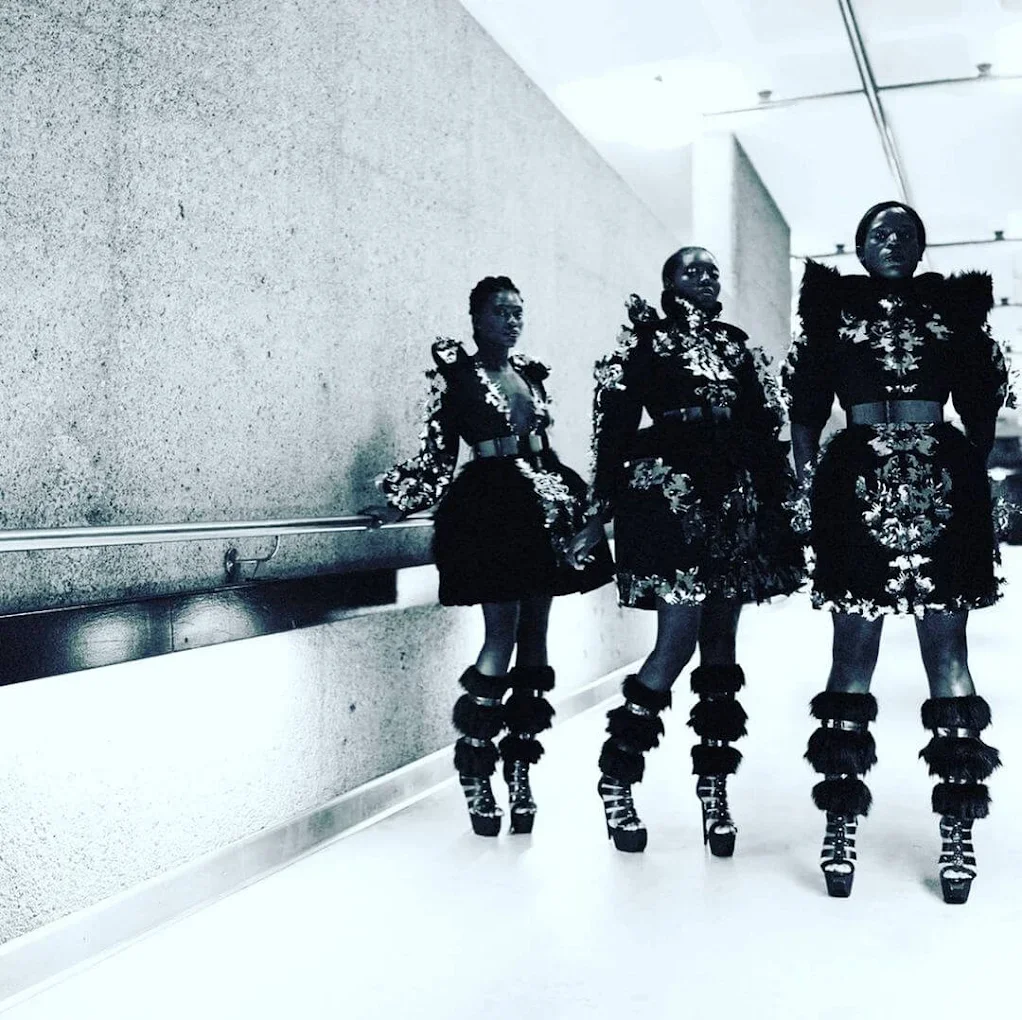 Three women walk in a corridor wearing matching black floral dresses, created for laser cut vinyl, with puffed shoulders and high-heeled platform shoes featuring fur-like embellishments.