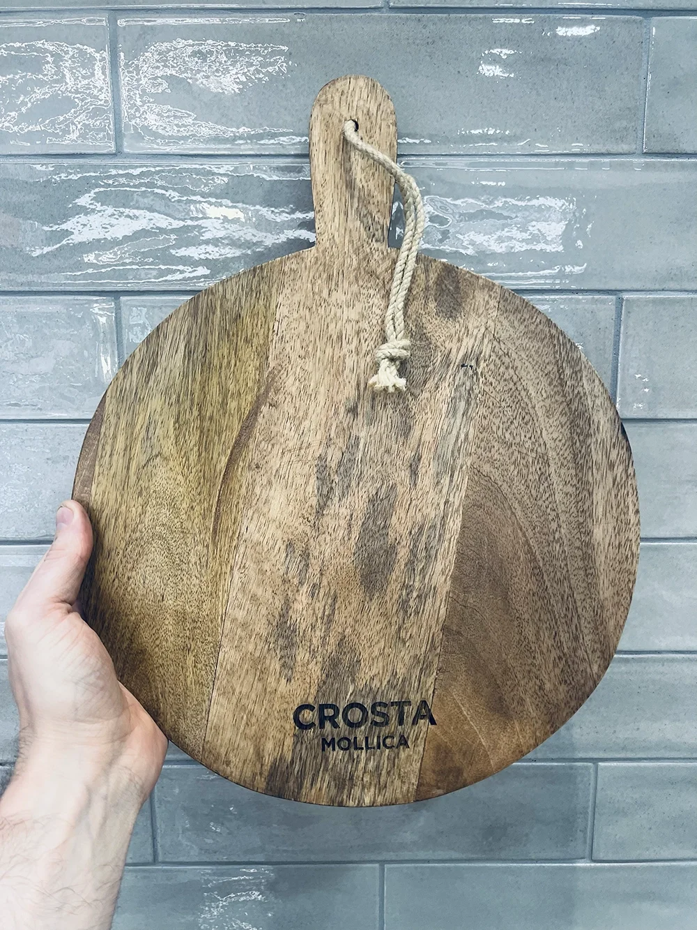 Round wooden cutting board with handle and rope loop, engraved with 'CROSTA MOLLICA', held by a hand against a grey tile wall background.
