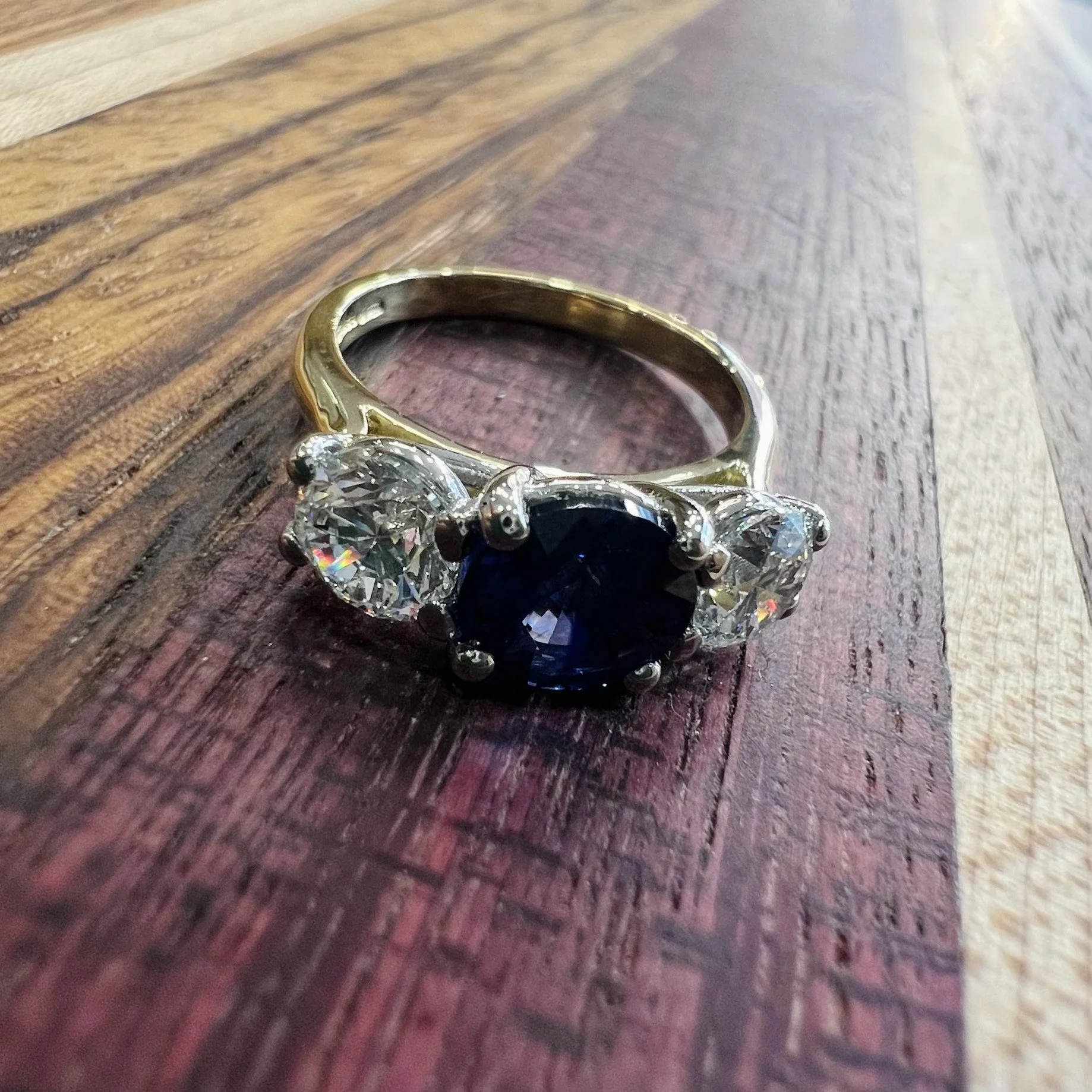 A trilogy engagement ring handcrafted in Hatton Garden. At its centre sits a deep, velvet-blue oval sapphire, flanked by two bright, round, brilliant-cut diamonds. Set in a polished yellow gold band with secure platinum claws