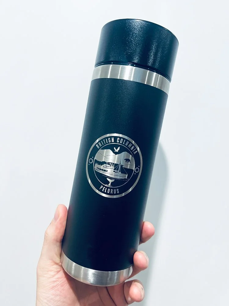 A hand holding a black travel thermos with a silver band and base, featuring an engraved circular logo of British Columbia Pelorus, which has sailboats, a whale, and trees.