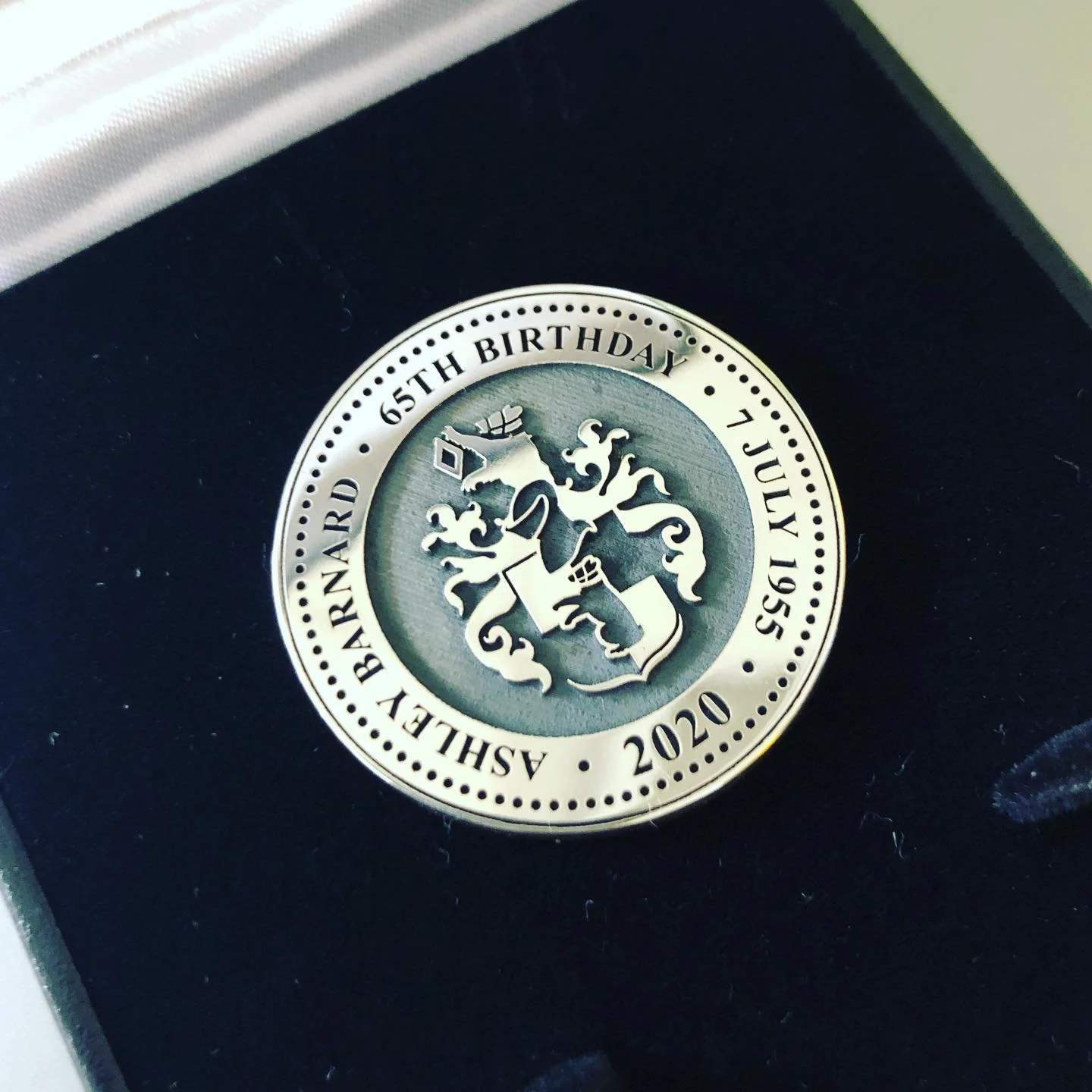 An engraved commemorative coin celebrating Ashlee Barnard’s 65th birthday on July 15, 2020, featuring a crest or emblem in the centre with a shield, a crown, and decorative elements, placed inside a black jewellery box.