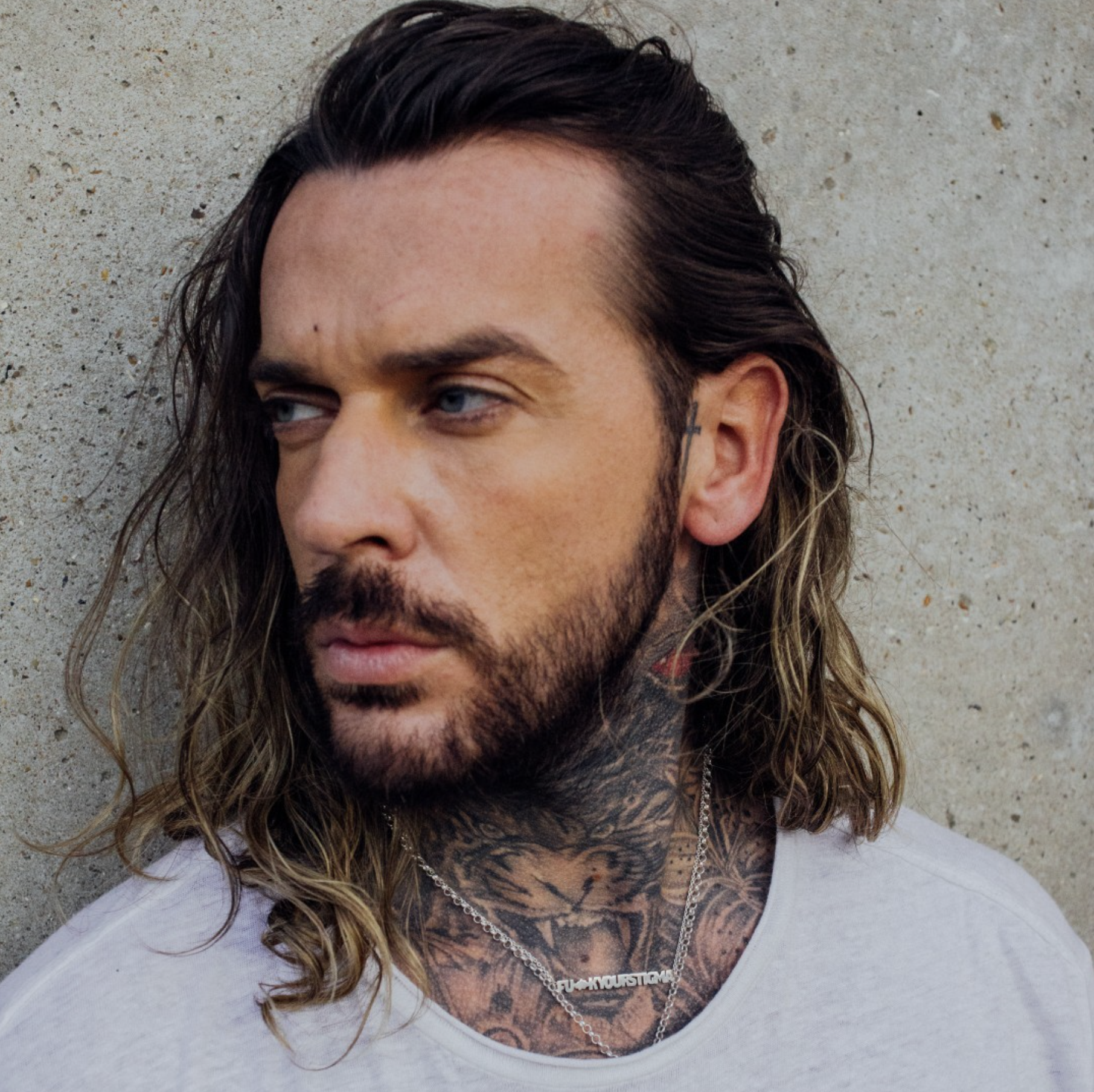 A man with long wavy hair, a beard, and tattoos on his neck and chest. He's wearing a white shirt and silver necklaces that have been laser cut, standing against a concrete wall.