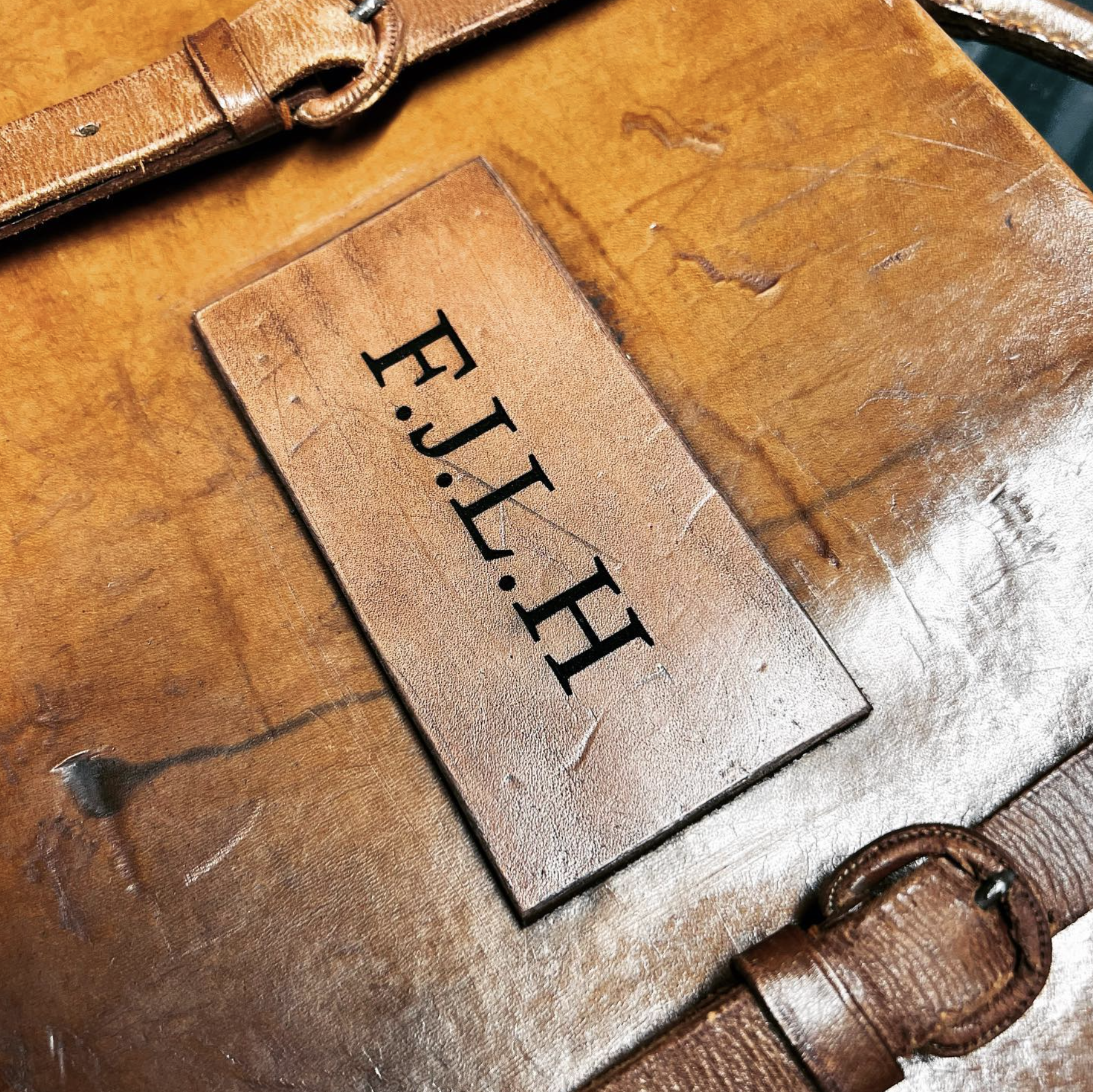 Close-up of a worn brown leather briefcase with a rectangular nameplate engraved with 'F.J.J. J.F' on its surface.