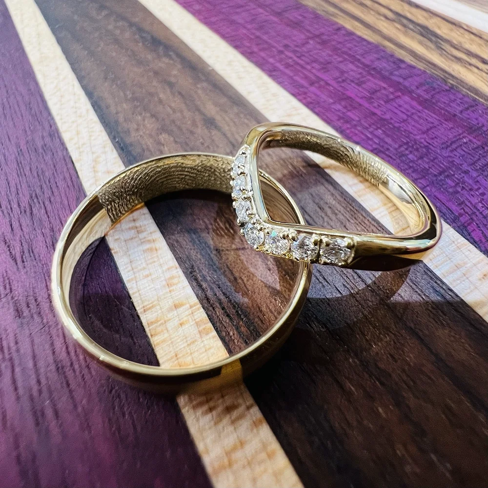 Engraved gold wedding rings, one plain and one with small diamonds, resting on a multicoloured wooden surface.