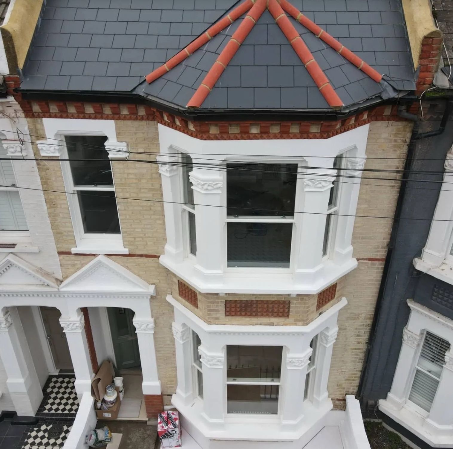 Aerial view of a three-story Victorian-style brick house with white decorative trim and bay windows. The roof is dark gray with red tiles stacked in a pattern on top.