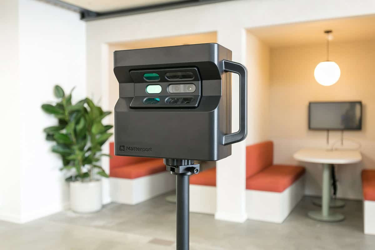 A Matterport 3D camera on a tripod inside a modern room with a tv, a round table, a built-in bench with red cushions, and a potted plant.