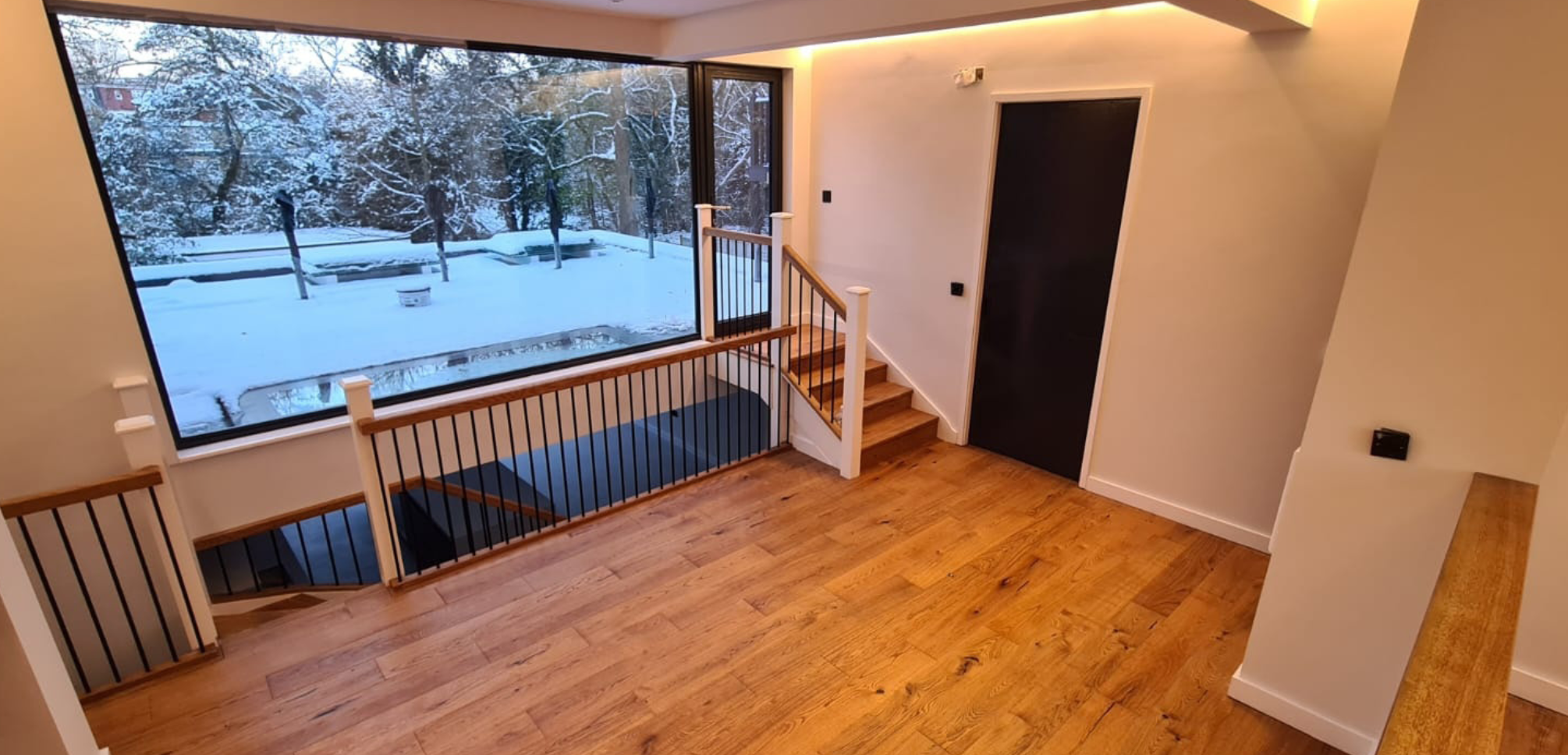 Interior view of a modern home with a large window showing a snowy outdoor landscape, including trees, a pathway, and benches. The room has wooden flooring, a staircase with wooden steps and black metal balusters, a black door, and minimal wall decor.