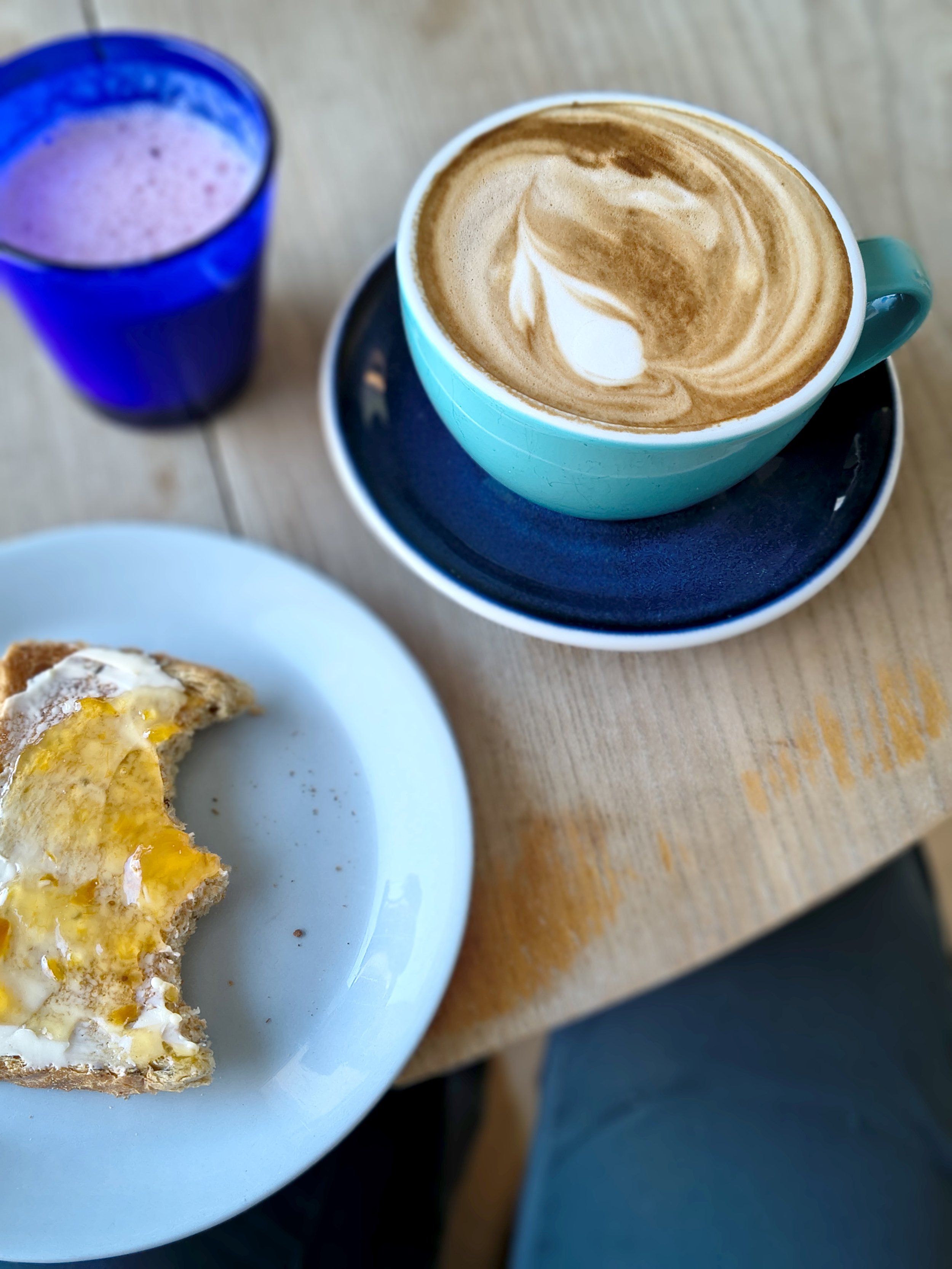 Kuvassa puisella pöydällä sinisellä lautasella puoliksi syöty rapea paahtoleipä appelsiinimarmeladilla, turkoosissa kupissa cappuccino kauniilla kuviolla ja sinisessä Kartio-lasissa marjasmoothie.
