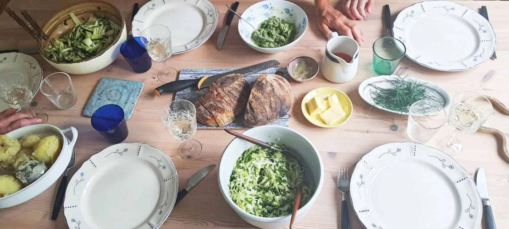 Ruoan ja juomien täyttämä ruokapöytä, jossa on leipää, salaatteja, perunoita, voita, lihaleikkeitä, juomia ja laseja.