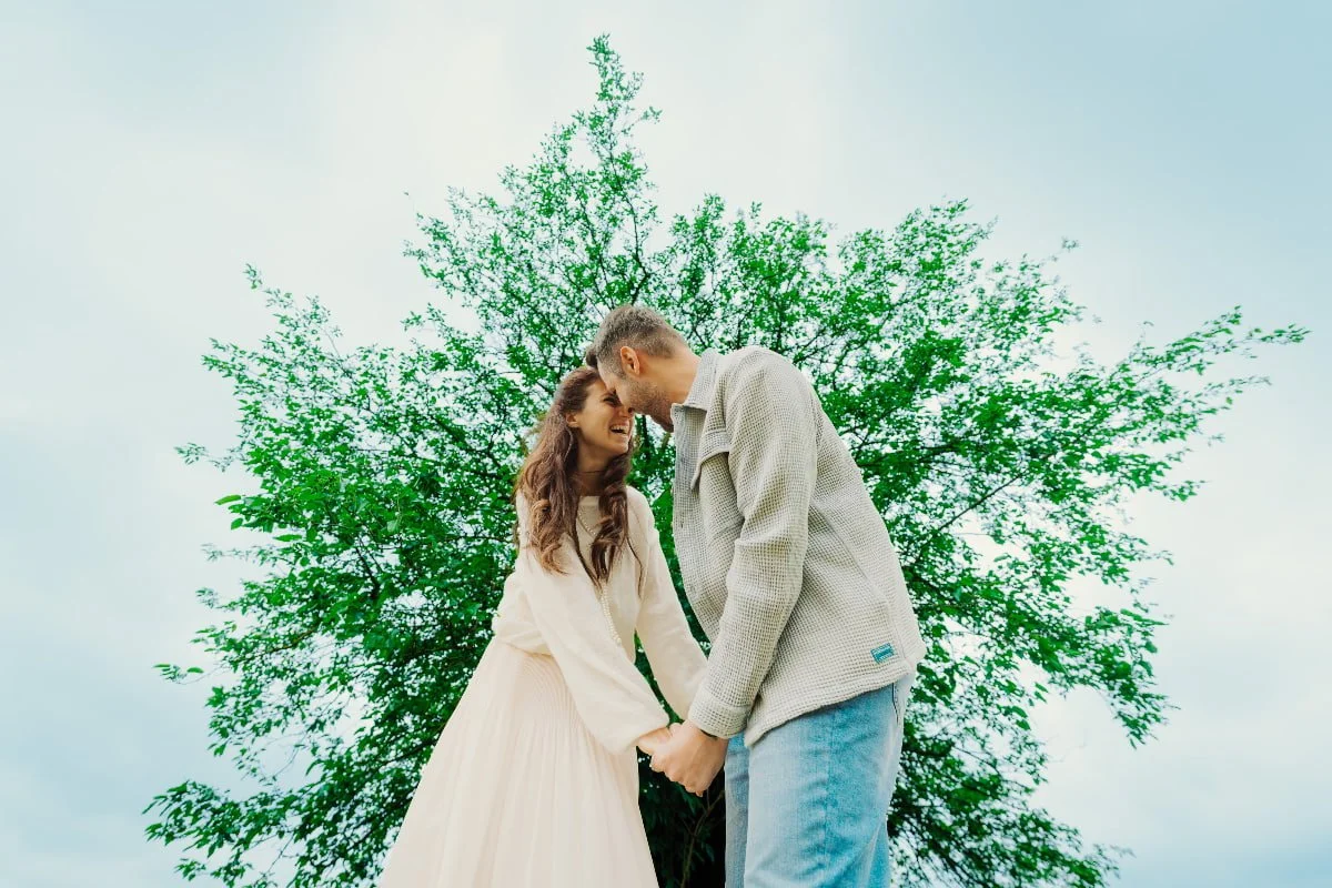 A couple holding hands and smiling in front of a large green-leafed tree on a cloudy day.
