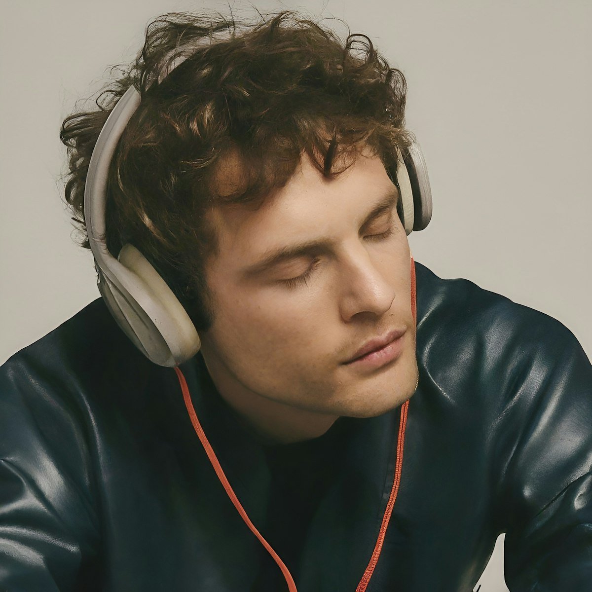 Young man with curly brown hair and light skin wearing large over-ear headphones and a black leather jacket, appears to be relaxed with eyes closed.