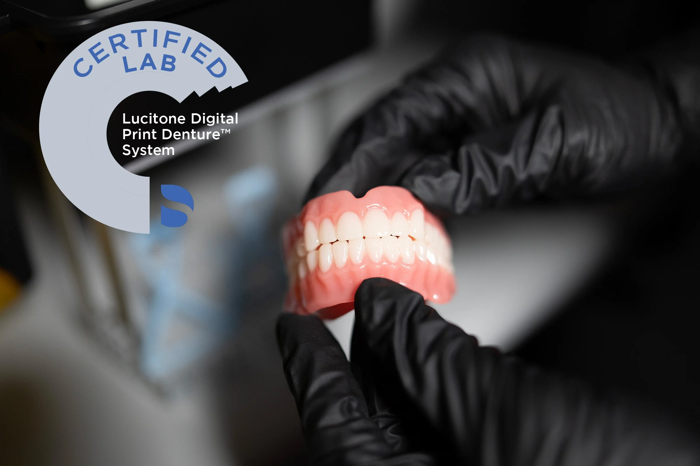 Close-up of a dental prosthetic denture being held by a hand in black gloves, with a logo on the upper left corner that reads 'CERTIFIED LAB Lucitone Digital Print Denture System'.