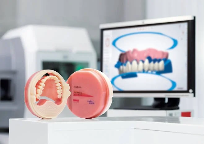 A dental model of teeth and gums, with a pink denture case, placed on a white surface in front of a computer monitor displaying a digital dental scan.