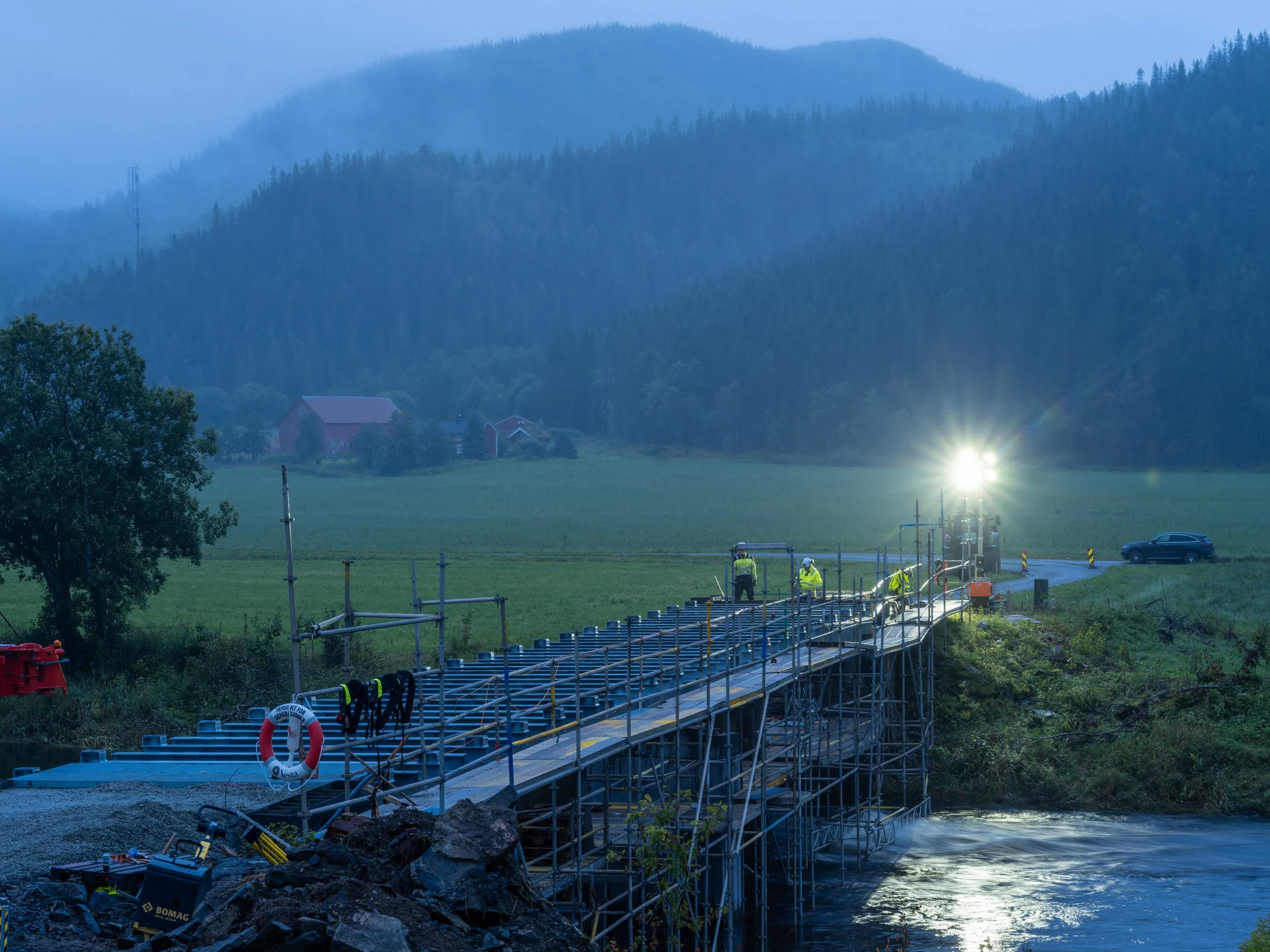 Breivold bru i Åfjord under anleggsarbeid i skumringen. Montører arbeider på stålkonstruksjonen, omgitt av stillaser, kraftige arbeidslys og elva nedenfor.