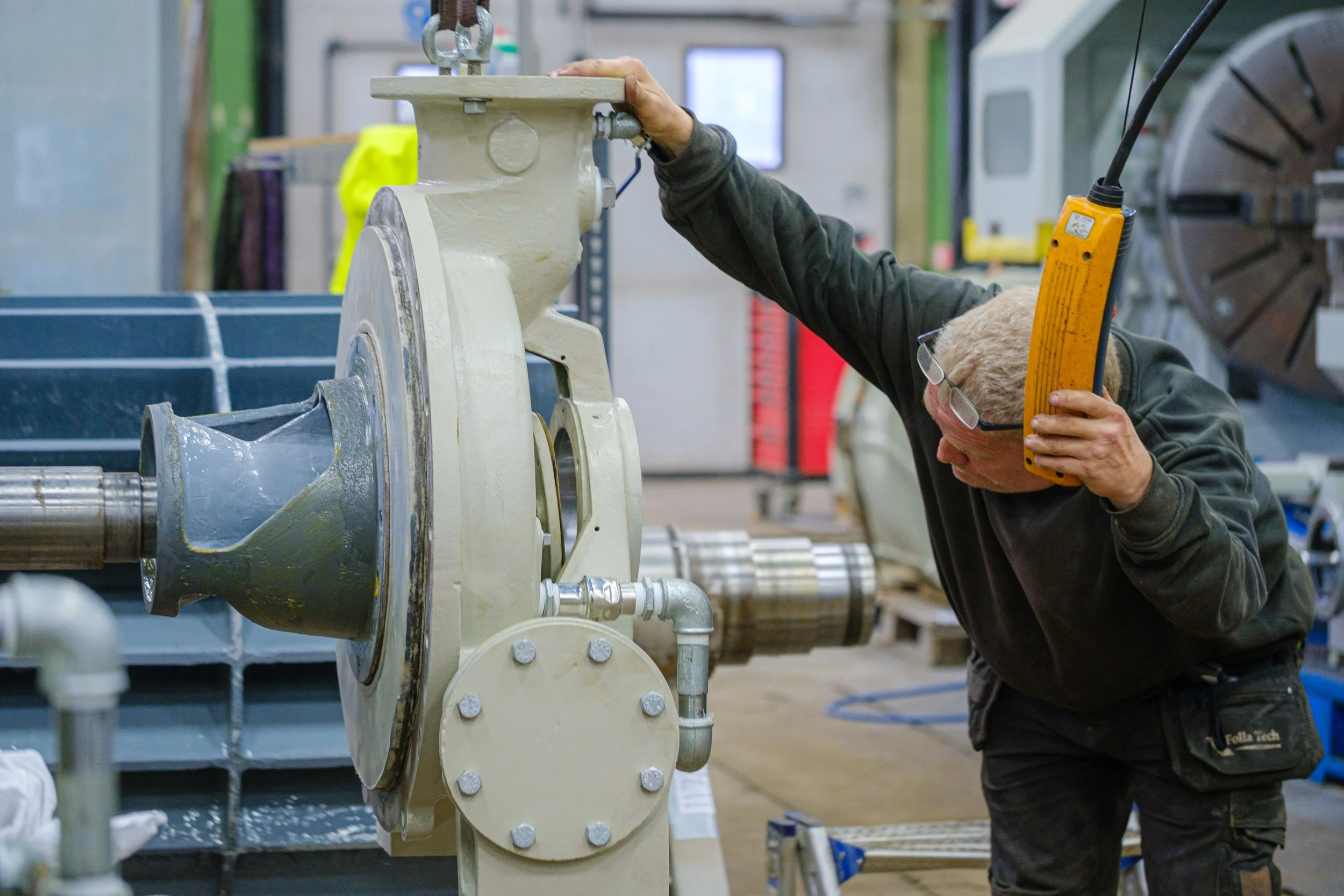 A man with safety glasses and a vest examining a large industrial machine part with a handheld device in a workshop.