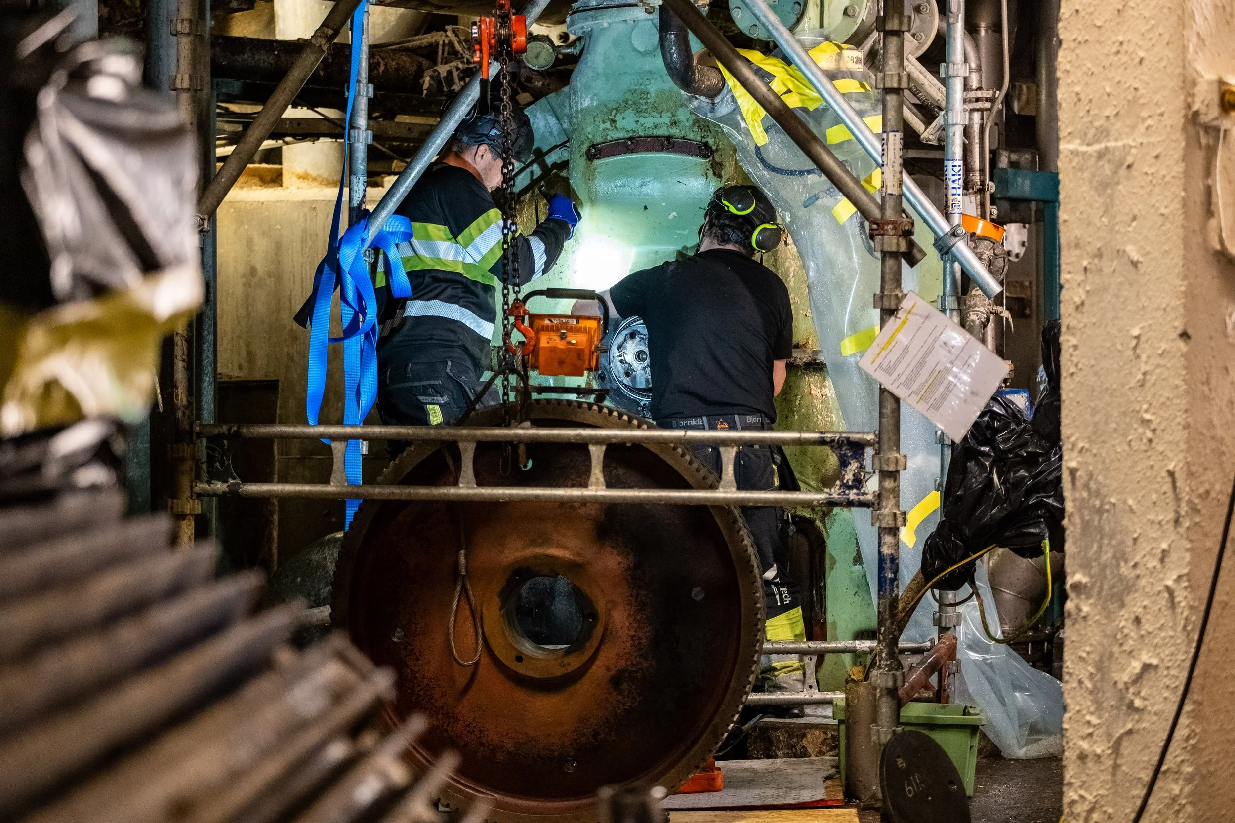 Two workers wearing protective gear, including ear protection and gloves, focusing on maintenance and repair work.