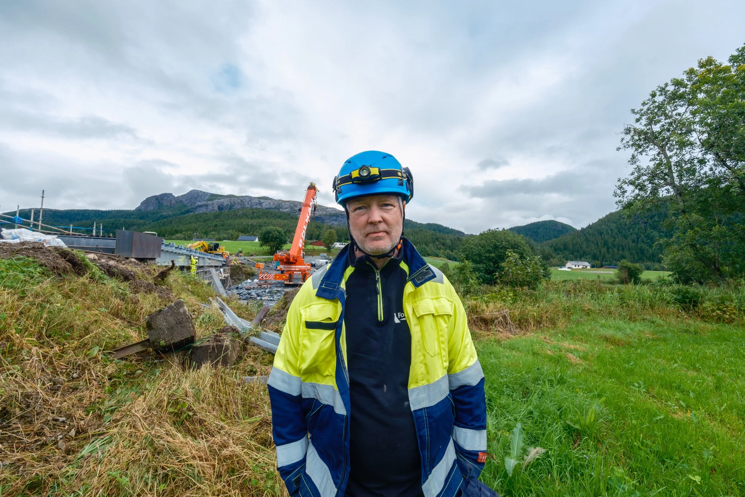 Terje Skjevik fra Folla Tech med blå vernehjelm og gul synlighetsjakke ved Breivold bru i Åfjord, med mobilkran og anleggsarbeid i bakgrunnen.