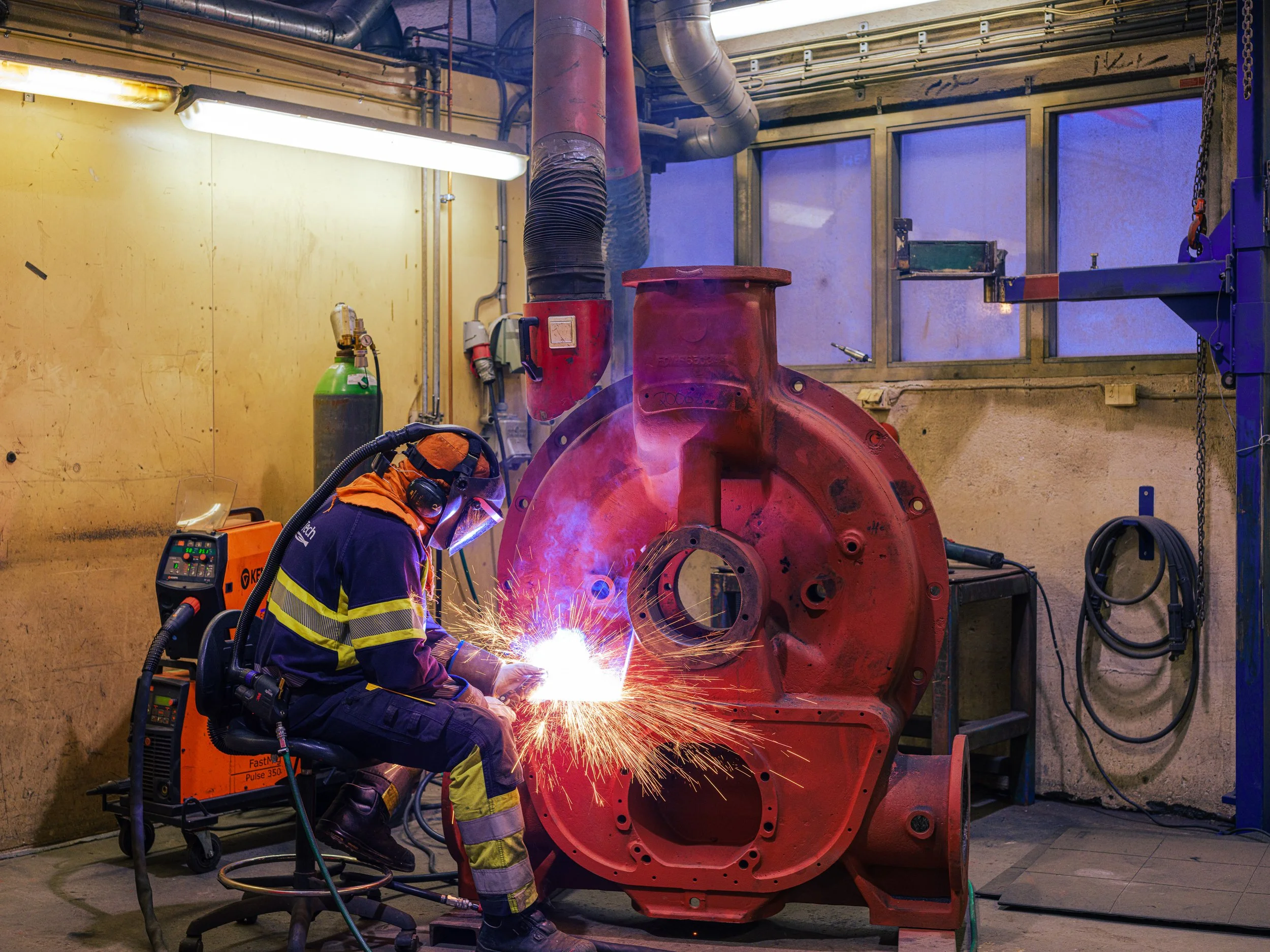 A worker wearing protective gear welds a large red industrial machine, with sparks flying in a workshop with yellow walls, pipes, and equipment.