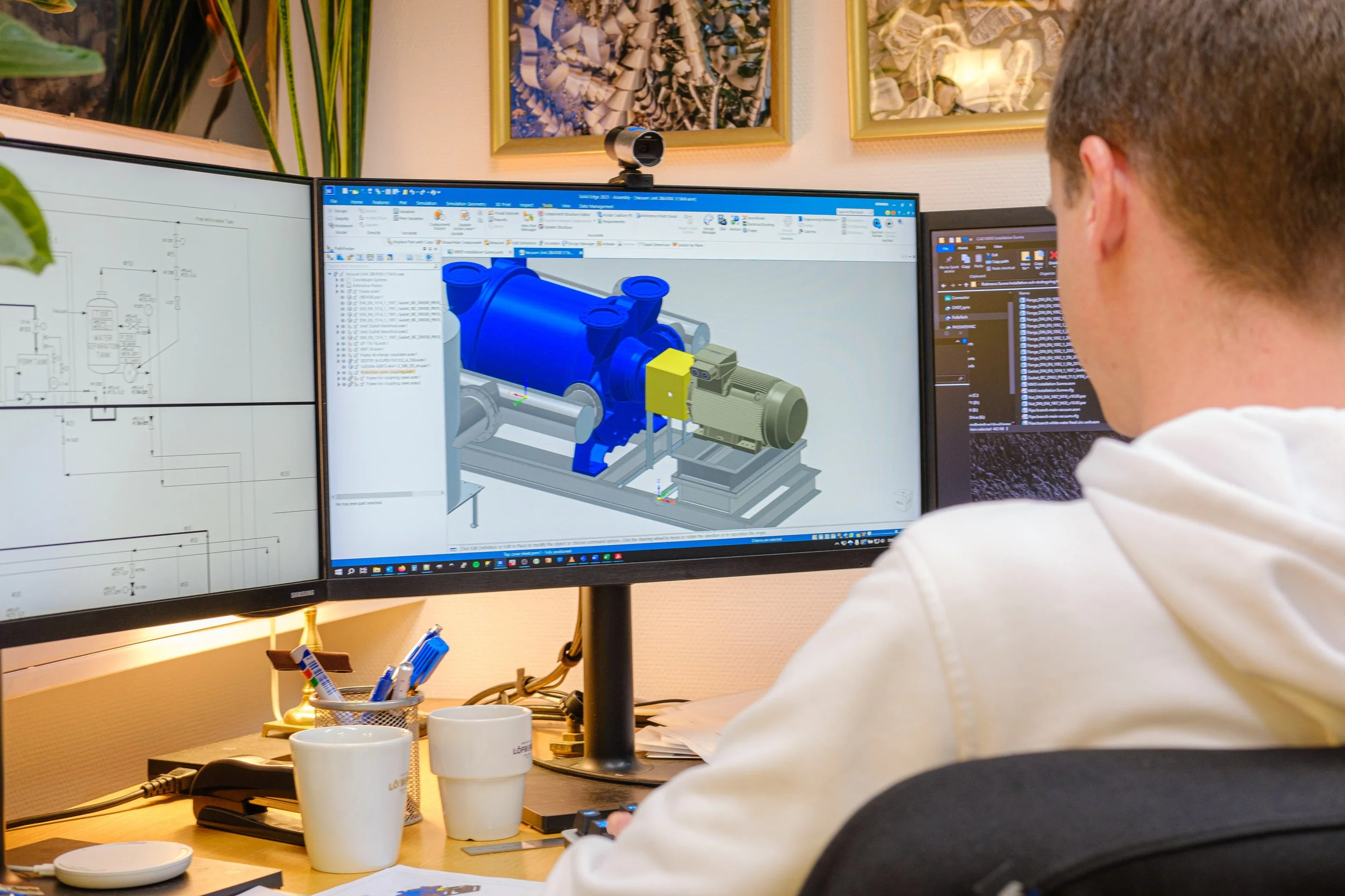 A person working at a desk with dual monitors, designing a 3D model of machinery on the right monitor and technical drawings on the left monitor, in an office environment.