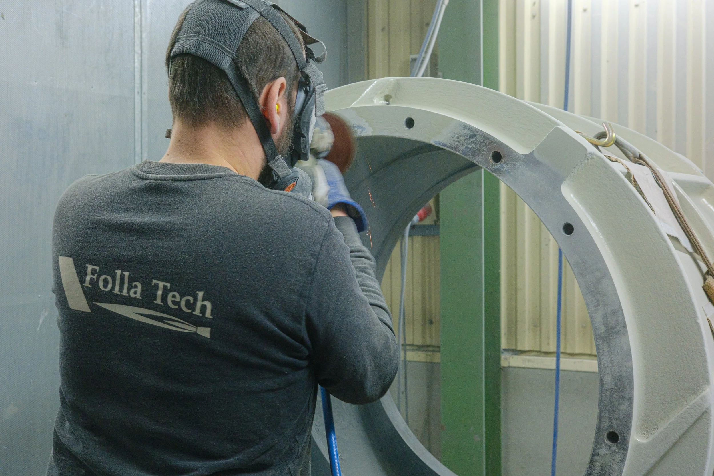 A worker wearing a Folla Tech branded shirt, helmet, and safety glasses, doing surface treatment on a pump in an industrial workshop.