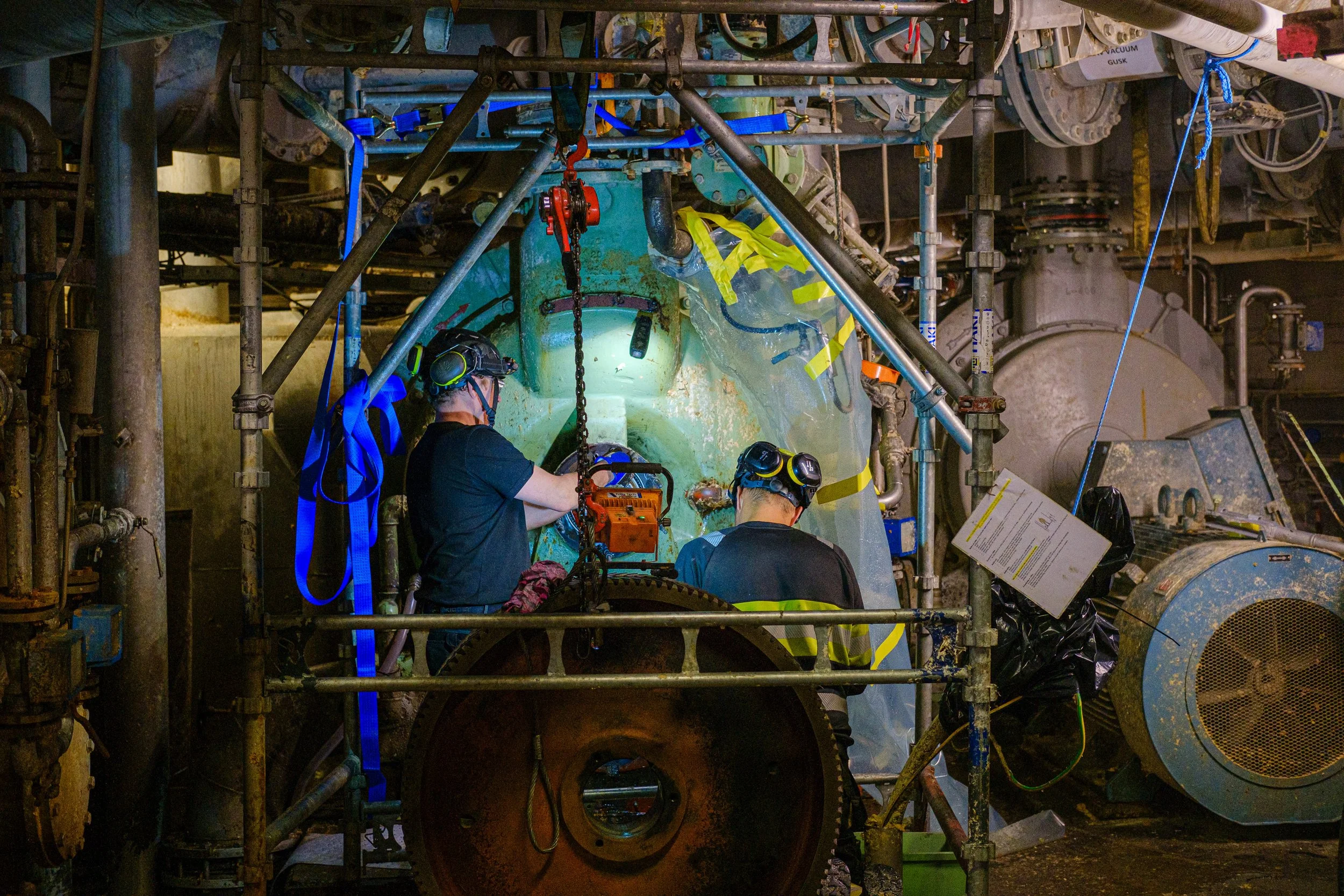 Two workers wearing protective gear working inside an industrial facility, surrounded by pipes, machinery, and scaffolding.