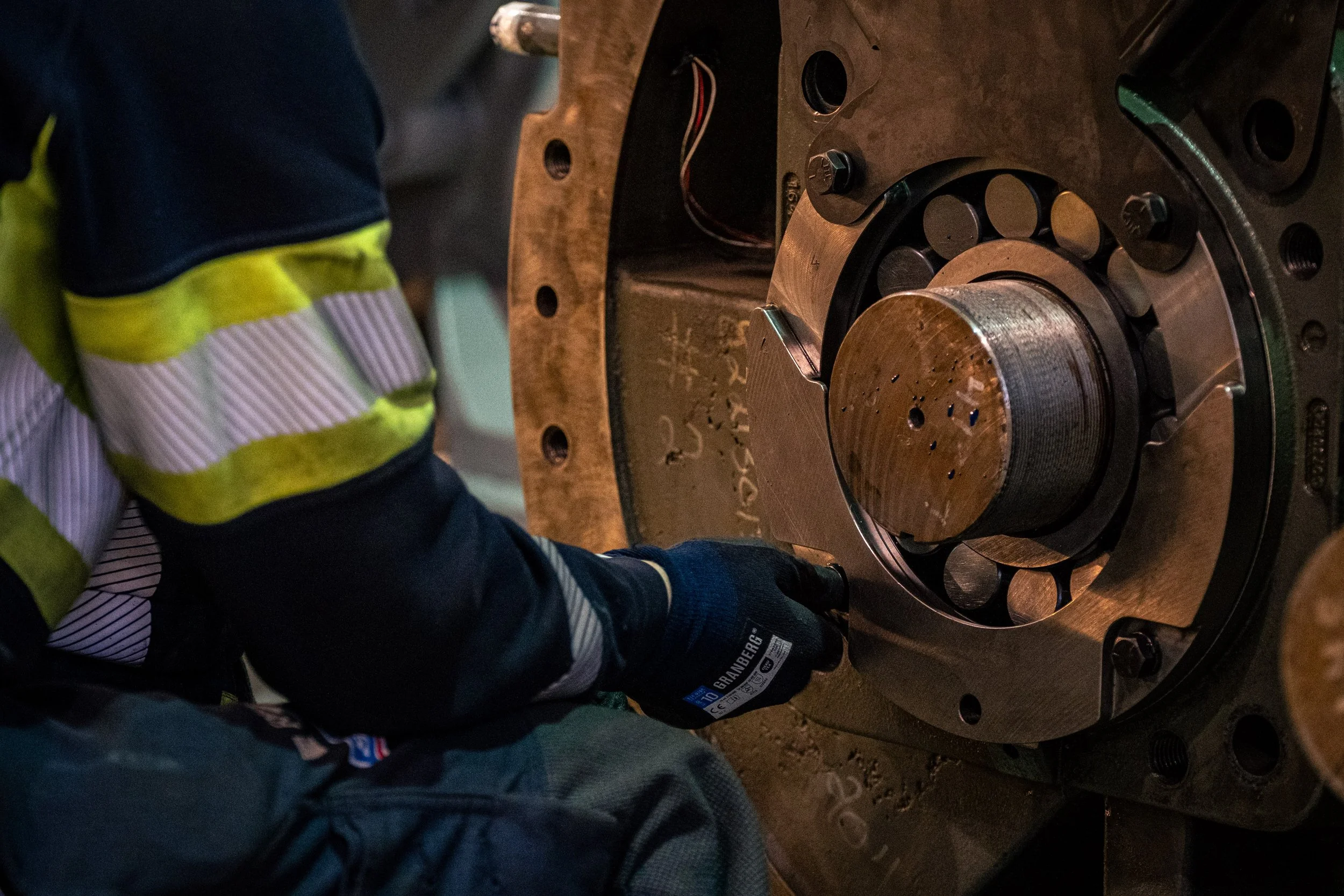 A worker in high visibility clothing and gloves is using a tool on a large, circular metal component, part of machinery or industrial equipment.