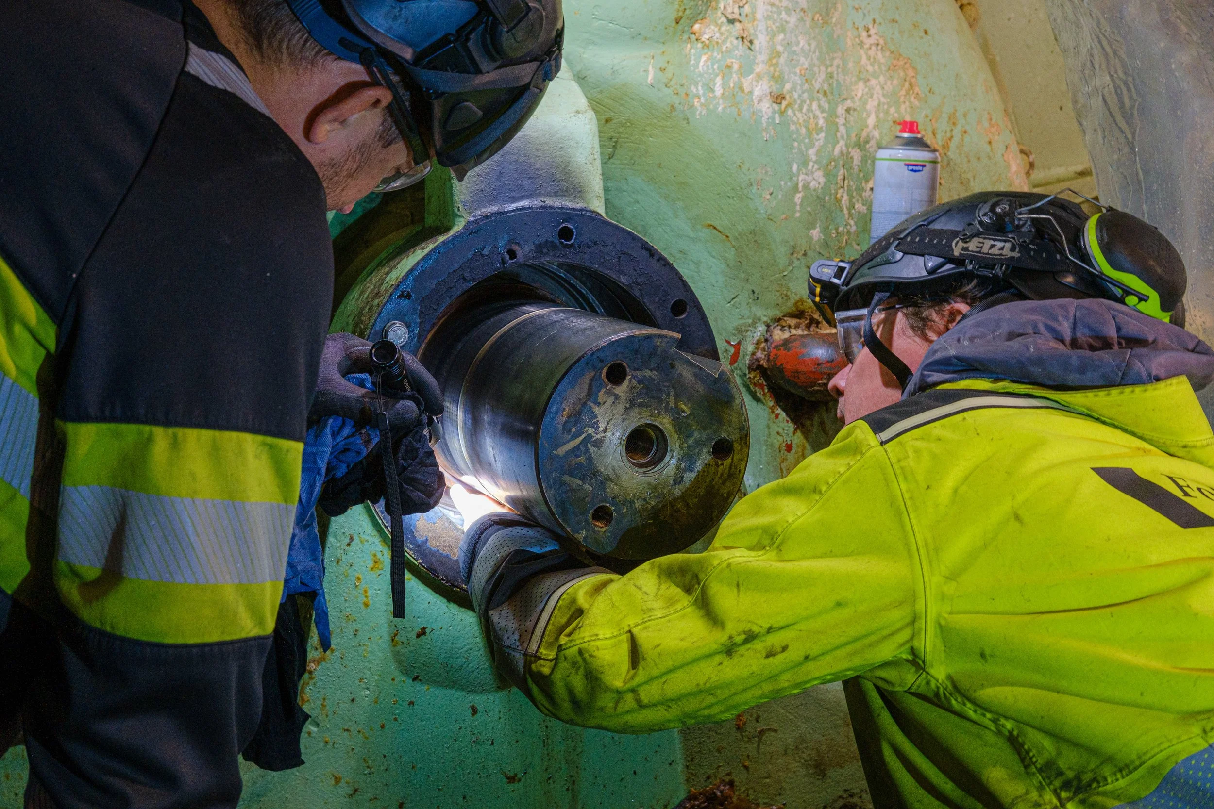 Two workers wearing helmets and protective gear are welding a large metal pipe in an underground tunnel or industrial setting.