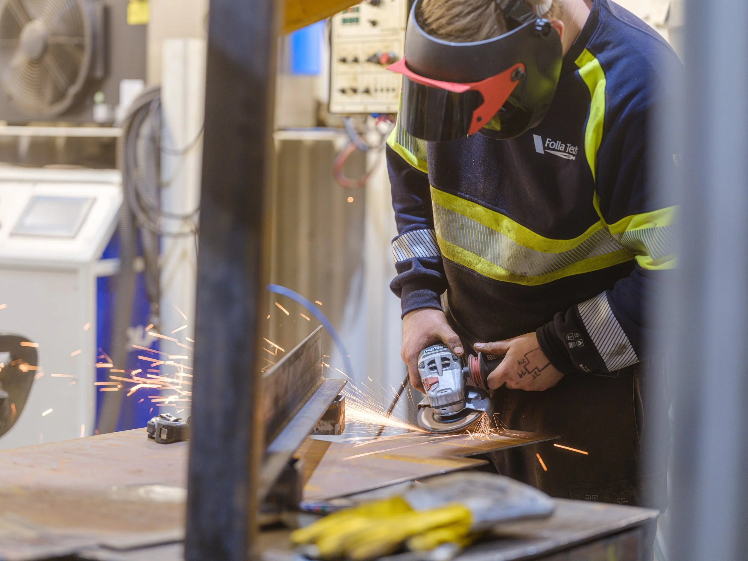 A worker in safety gear uses an angle grinder on a piece of metal, producing sparks in a workshop or industrial setting.