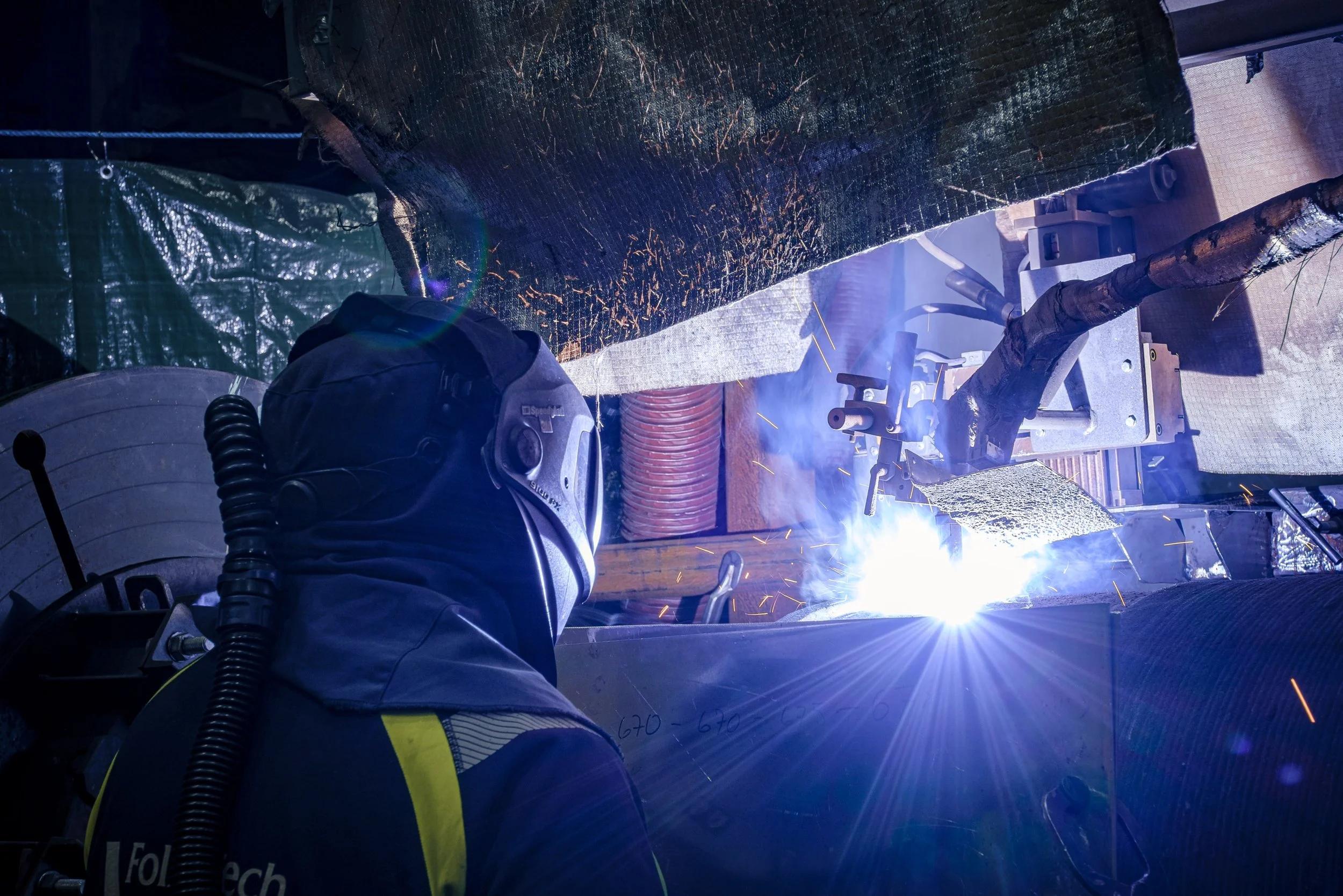 A welder in protective gear welding metal, with sparks and bright light and smoke showing in an industrial workshop.