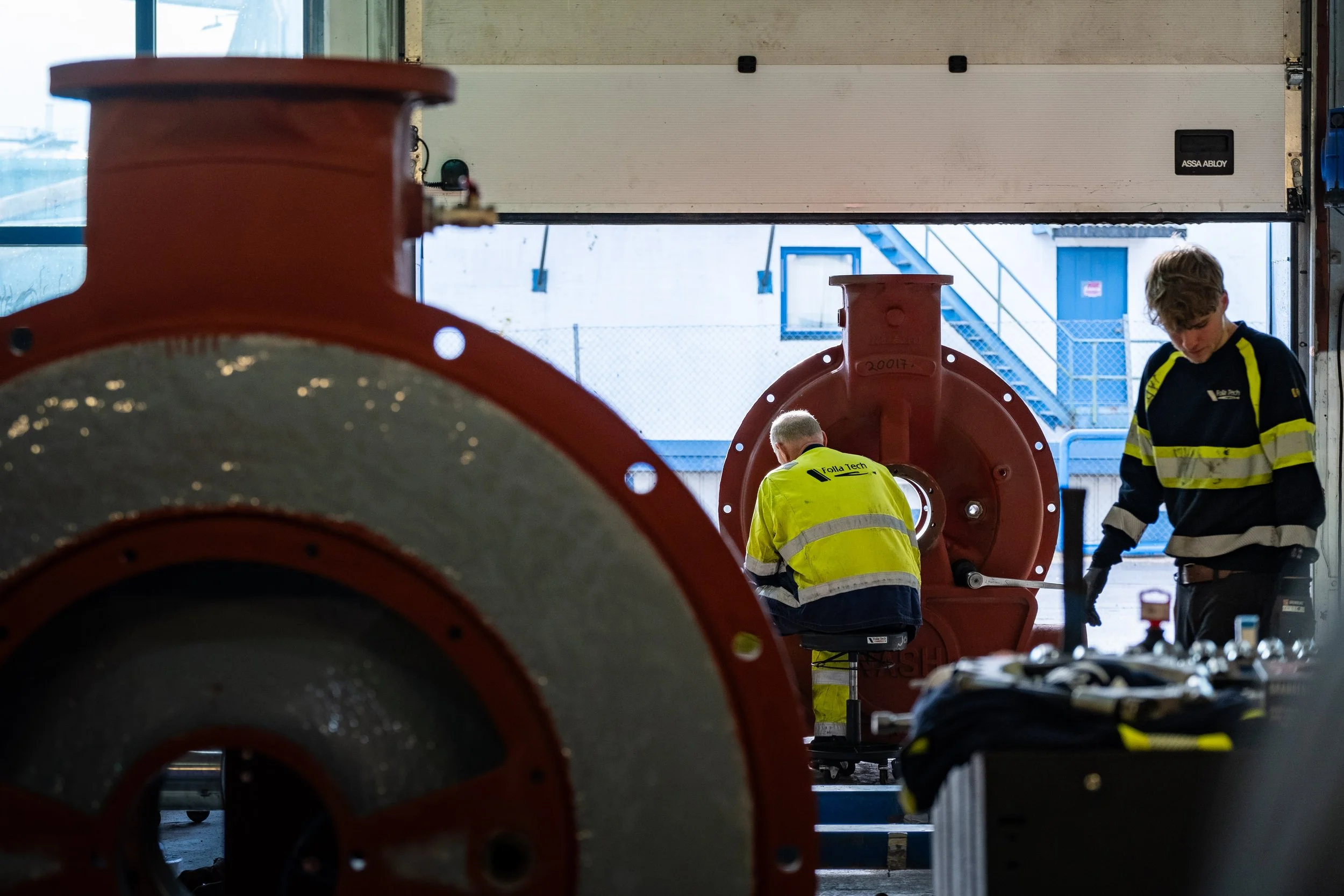 Two workers working on large industrial equipment or machinery. One worker is examining the component, while the other stands nearby, holding a tool.