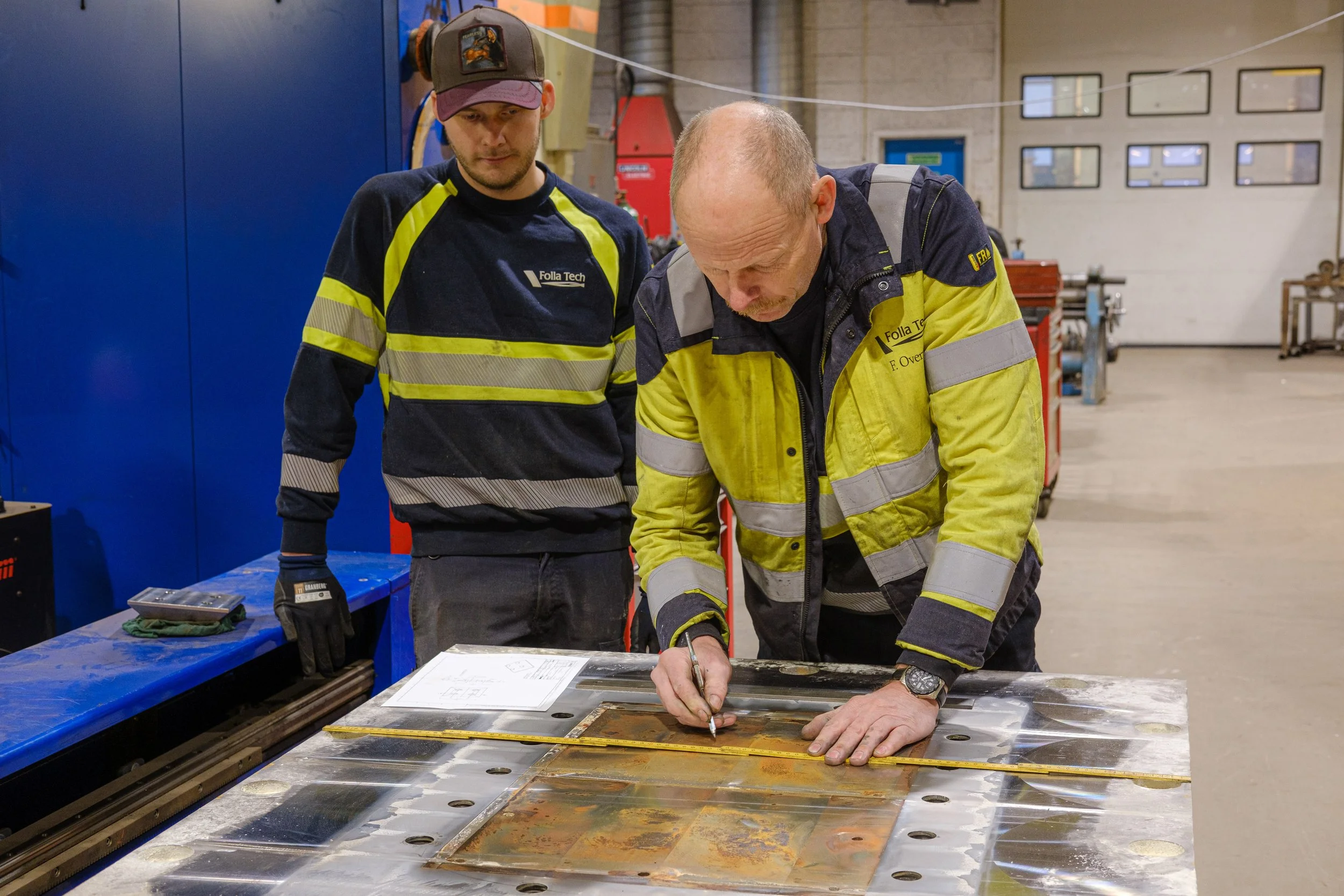 Two workers in safety gear examining a metal sheet with technical drawings and measuring tools in an industrial workshop.