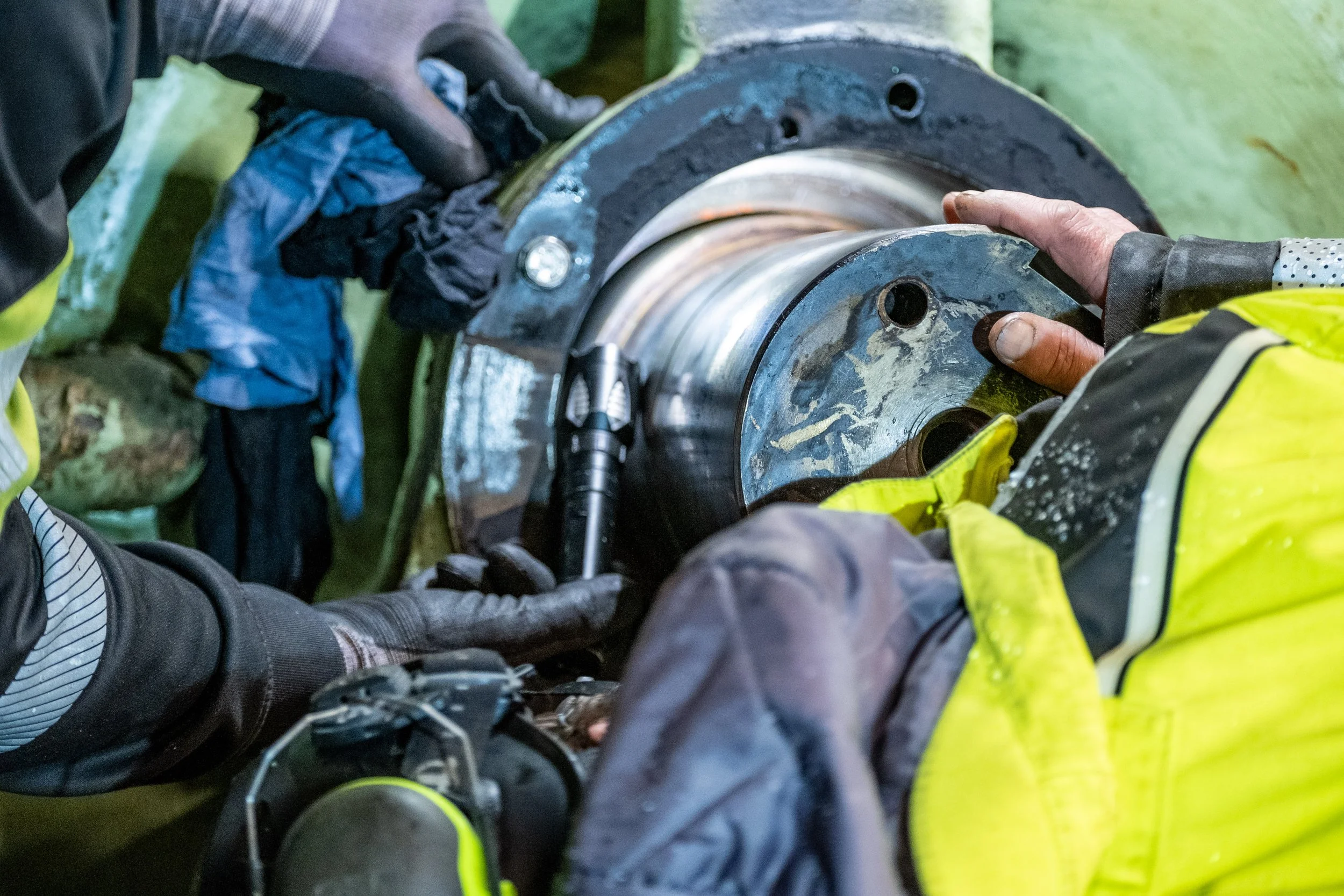 Two workers in high-visibility jackets performing maintenance on a large mechanical component using tools.