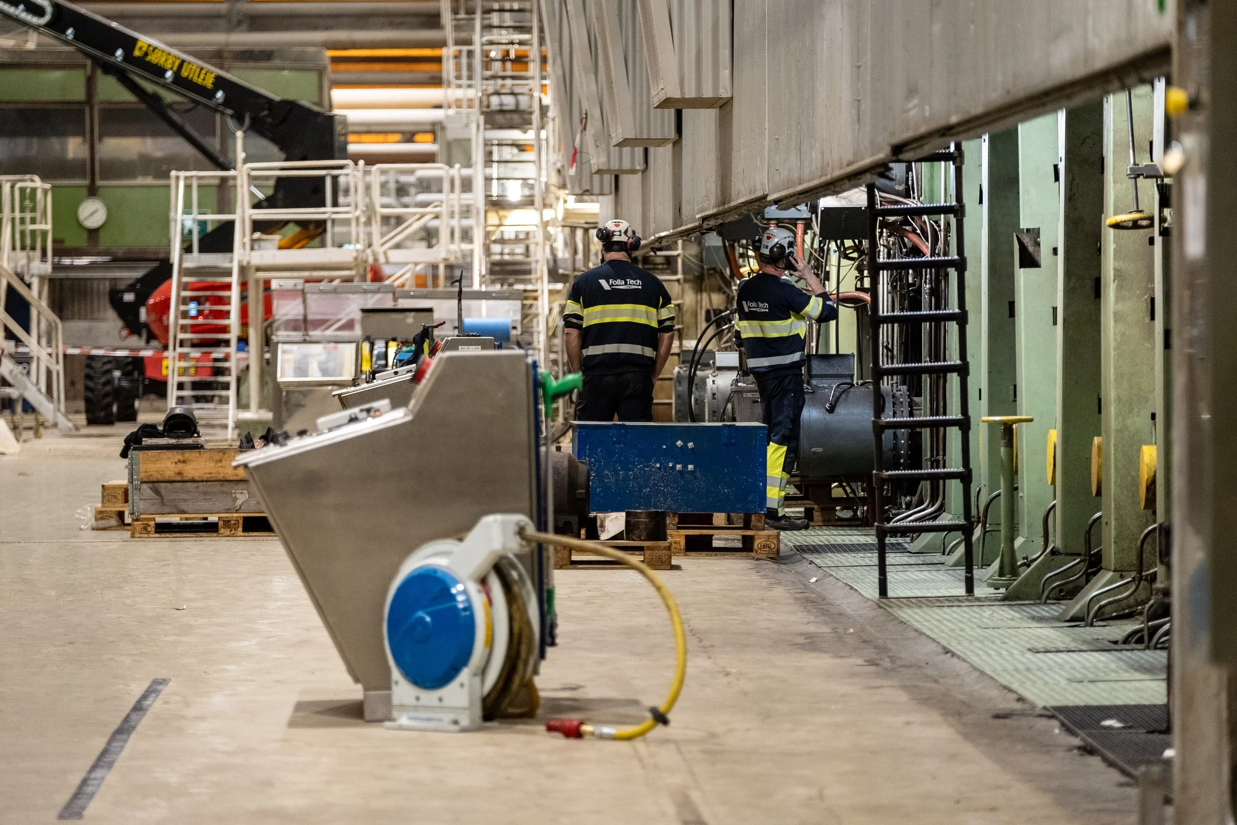 Two workers in safety uniforms and helmets working in an industrial facility, with machinery and equipment around them.