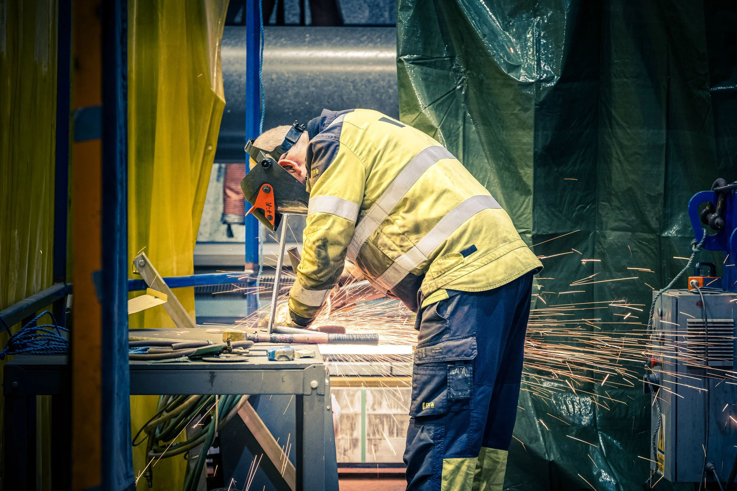 Worker in protective gear welding metal with sparks flying, inside a workshop surrounded by colored safety curtains.
