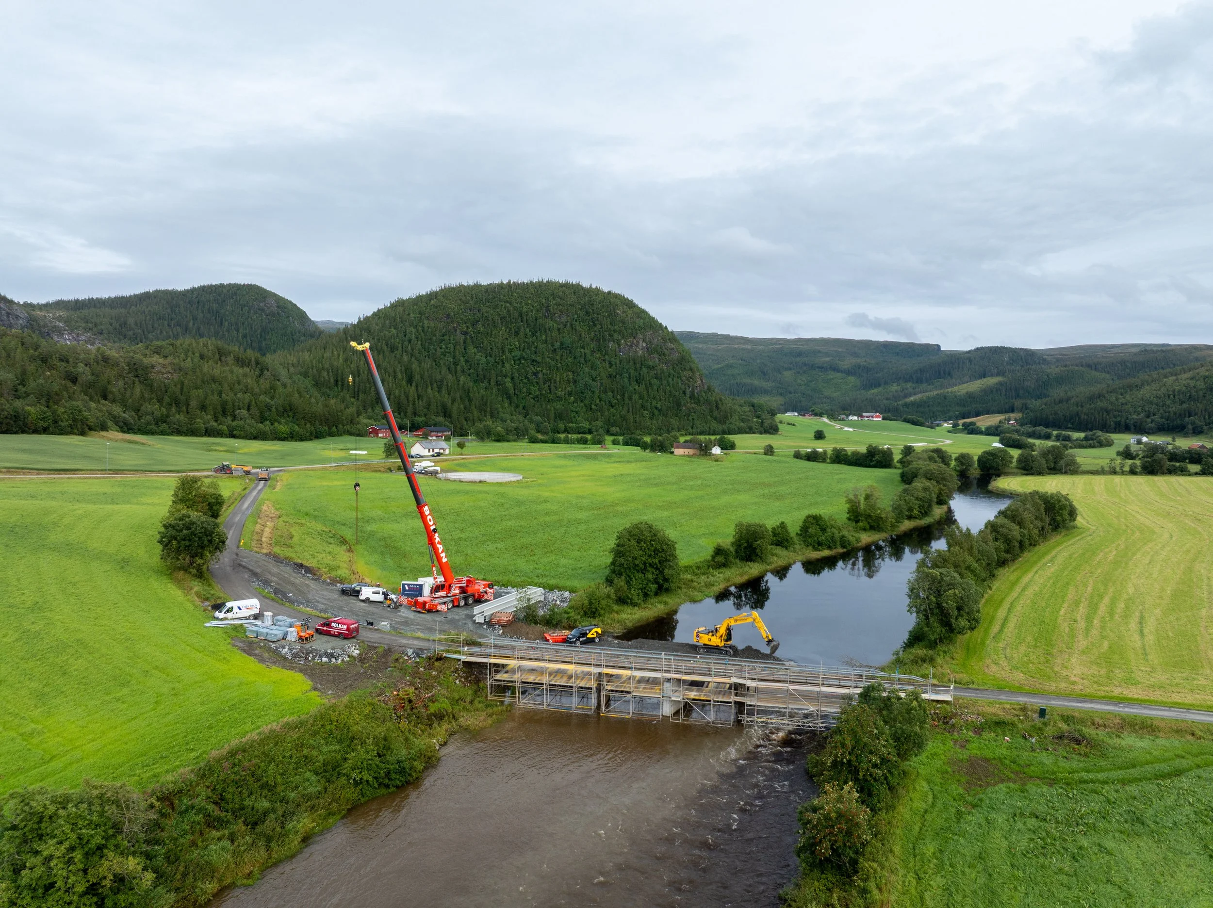 Dronefoto av Breivold bru i Åfjord under anleggsarbeidene, med stor mobilkran, gravemaskiner og anleggsområde omgitt av elv, jordbrukslandskap og skogkledde åser.