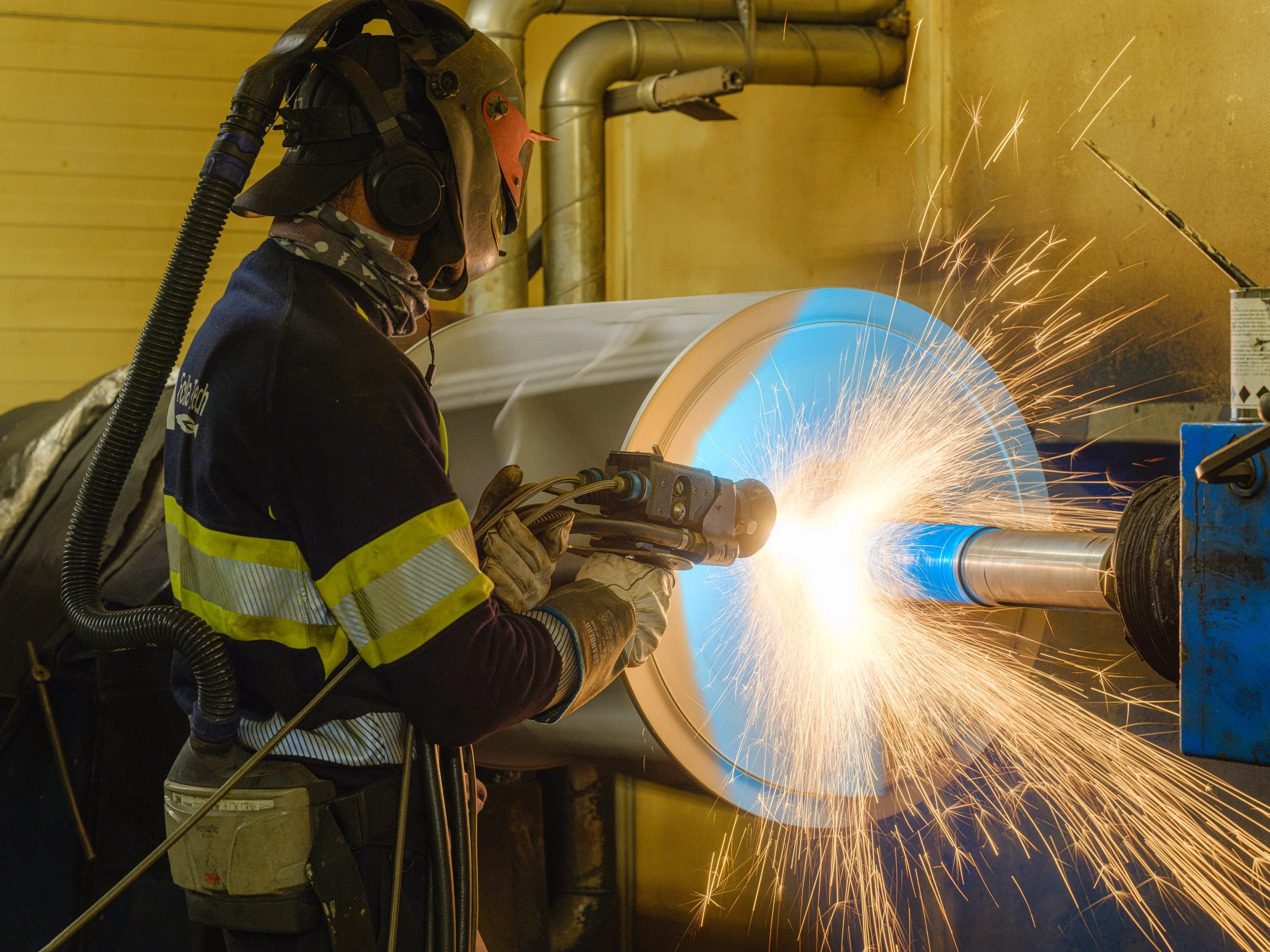 A worker using a welding tool on a large pipe, with sparks flying in an industrial setting.