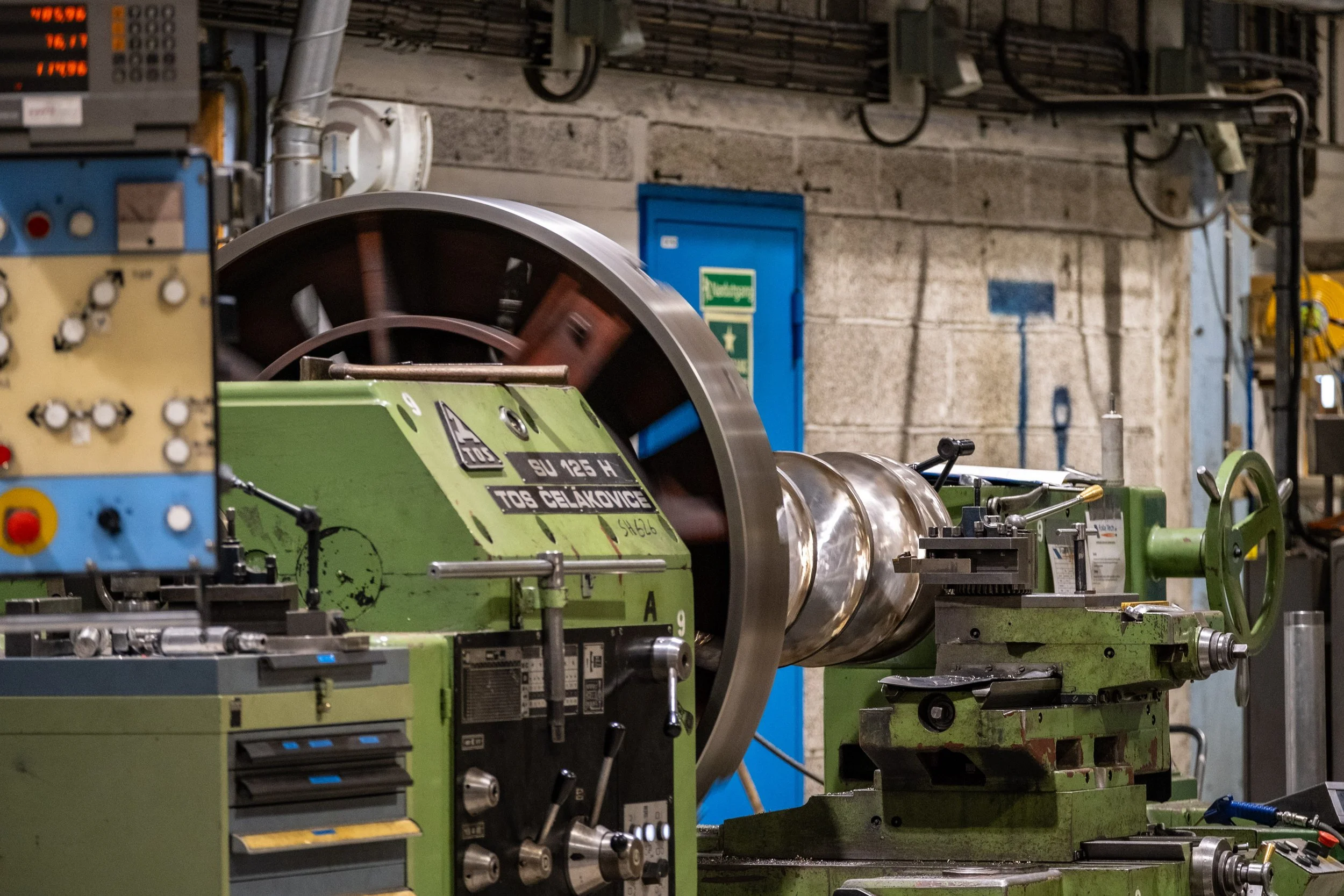 Industrial workshop featuring a large green metal lathe machine, with various controls and gauges, set against a concrete wall with electrical panels and wiring.
