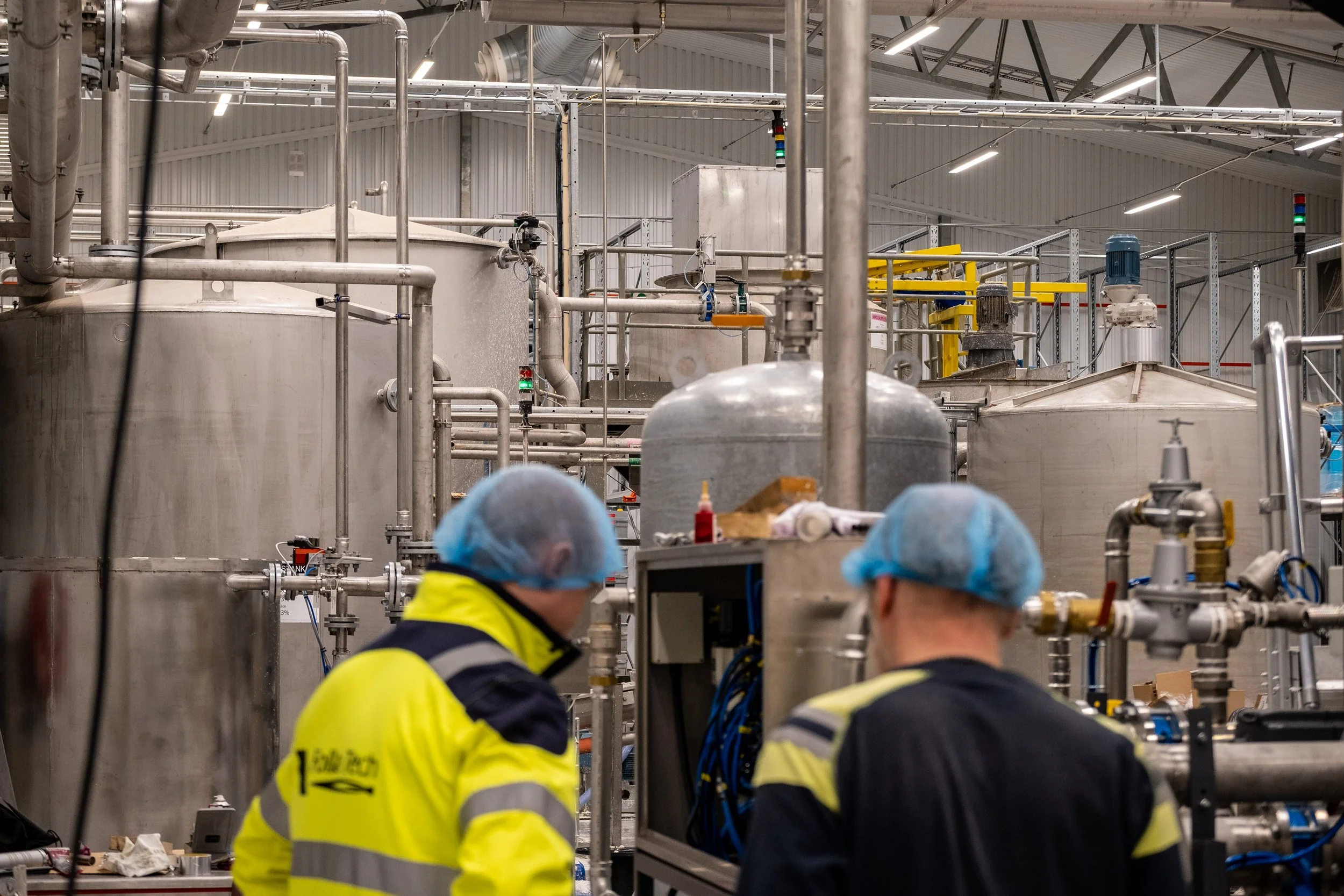 Two workers wearing yellow and black jackets, blue hairnets, and safety glasses working in a large industrial process plant with stainless steel tanks and piping.