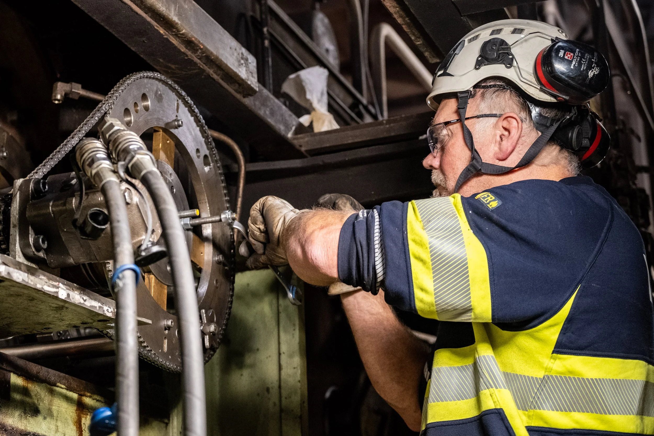 A man wearing safety glasses, helmet with hearing protection, a navy and yellow high-visibility shirt, and work gloves is working on a large mechanical component with pipes and gears.