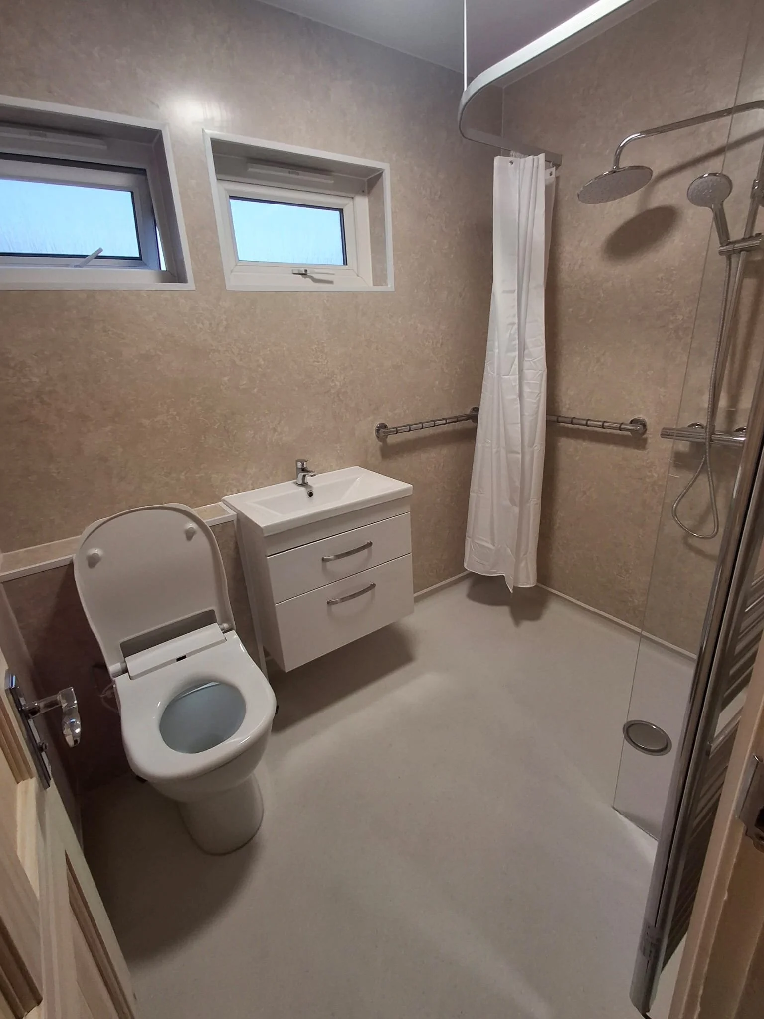 A bathroom with two small windows, a toilet with the lid open, a white sink with two drawers below, and a walk-in shower enclosed with a tiny shower curtain, with beige walls and a white floor.
