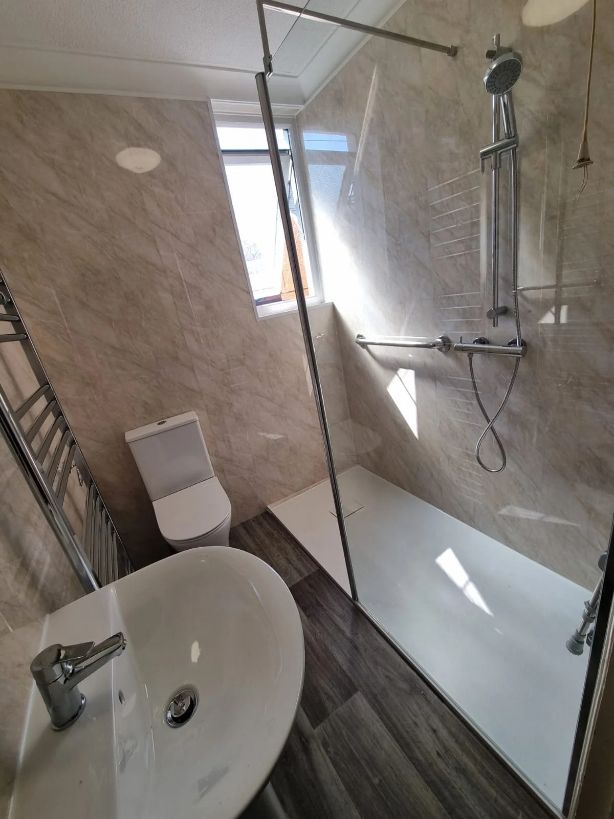 Modern bathroom with a walk-in shower, toilet, and sink.:The shower has a glass partition, chrome fixtures, and a window allowing natural light. The wall tiles are beige marble, and the floor is dark wood-look tile.