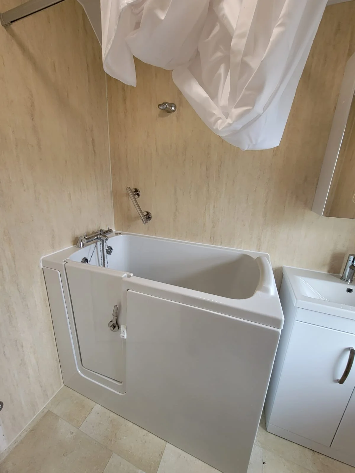Hospital bathroom with a white bathtub fitted with a door for wheelchair access, a small sink, and beige tiled walls and floor.