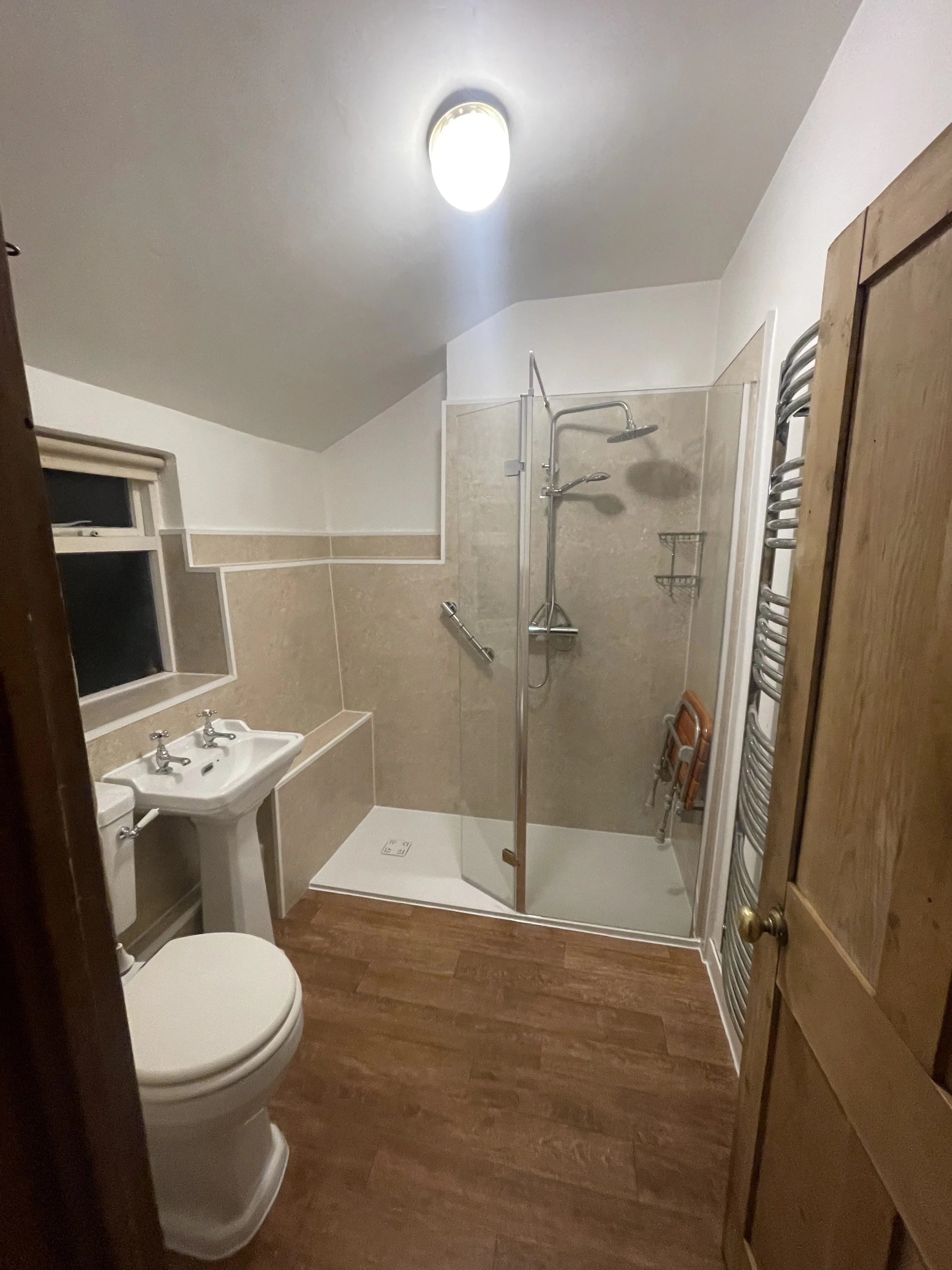 A small bathroom with a toilet, a twin sink, and a walk-in shower with a glass door. The bathroom has wood flooring, a small window, and a ceiling light.