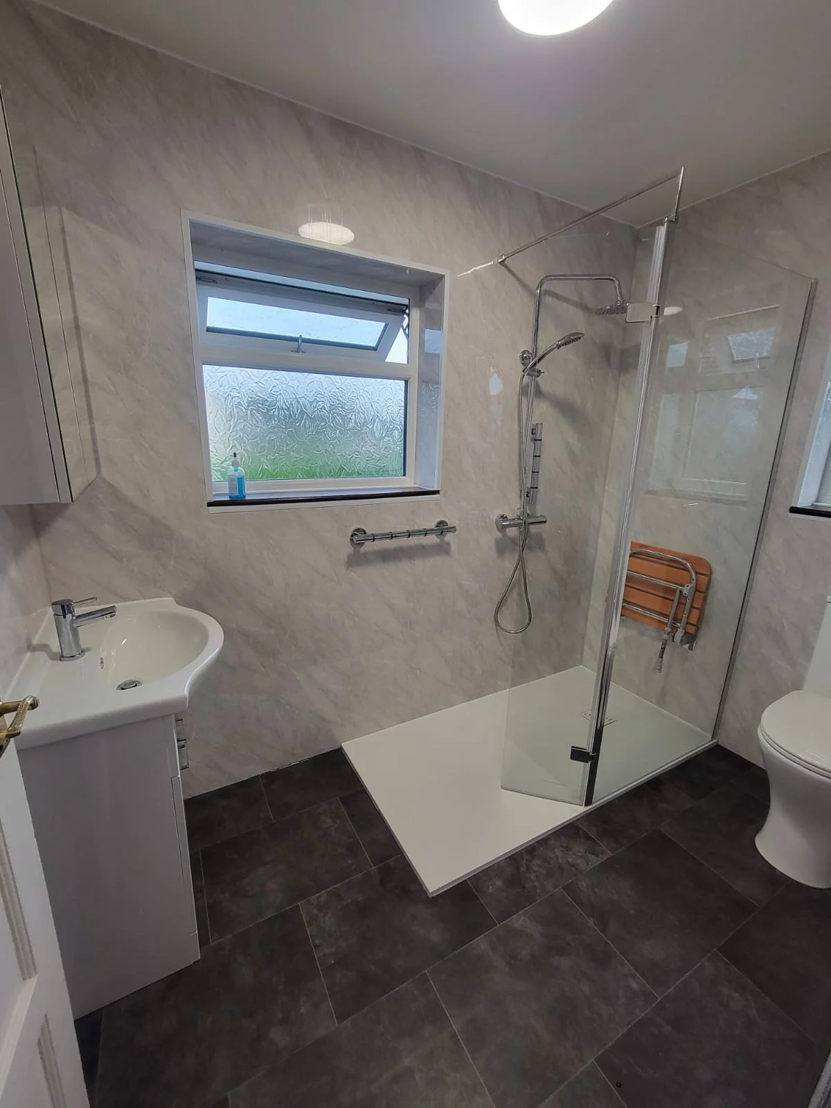 Modern bathroom with walk-in shower, small white sink, toilet, frosted window, dark tile flooring, and light-colored wall tiles.