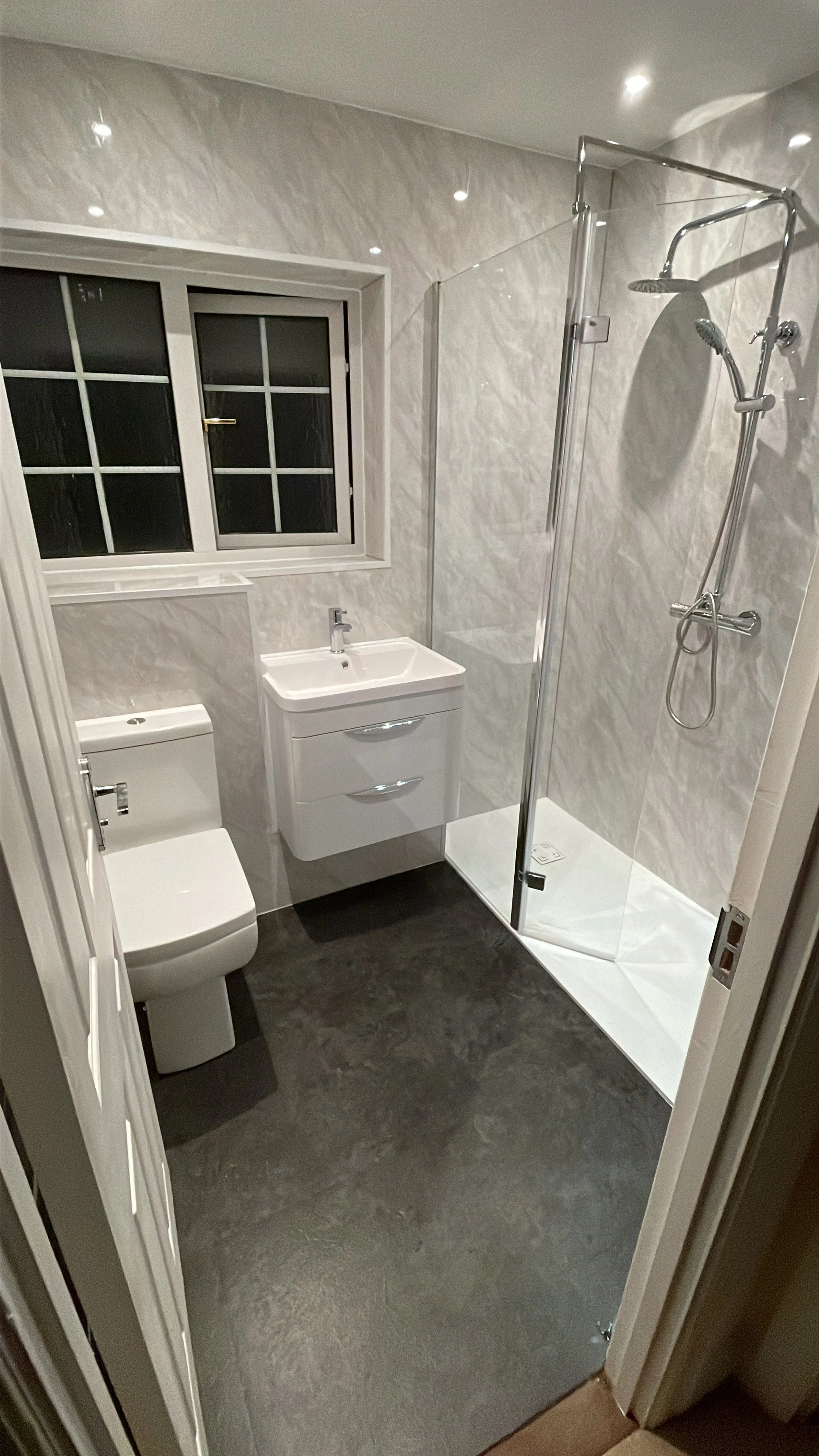 A modern bathroom with a white toilet, a small white vanity with a sink, a window with multiple panes, and a glass-enclosed shower with a rainfall and handheld showerhead.
