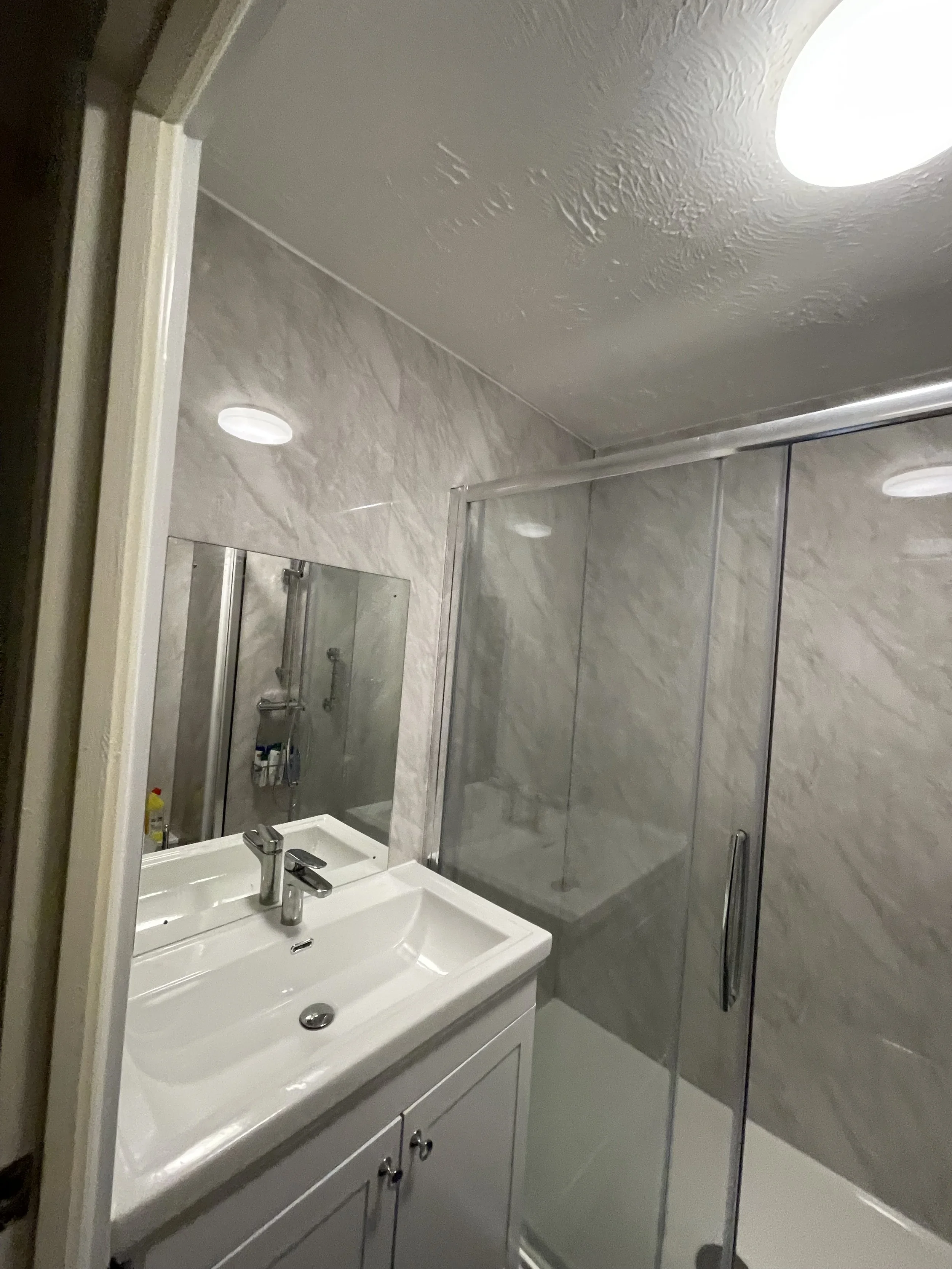 A small bathroom with a white vanity sink, a mirror mounted on beige tiled wall, a shower with glass door, and a ceiling light