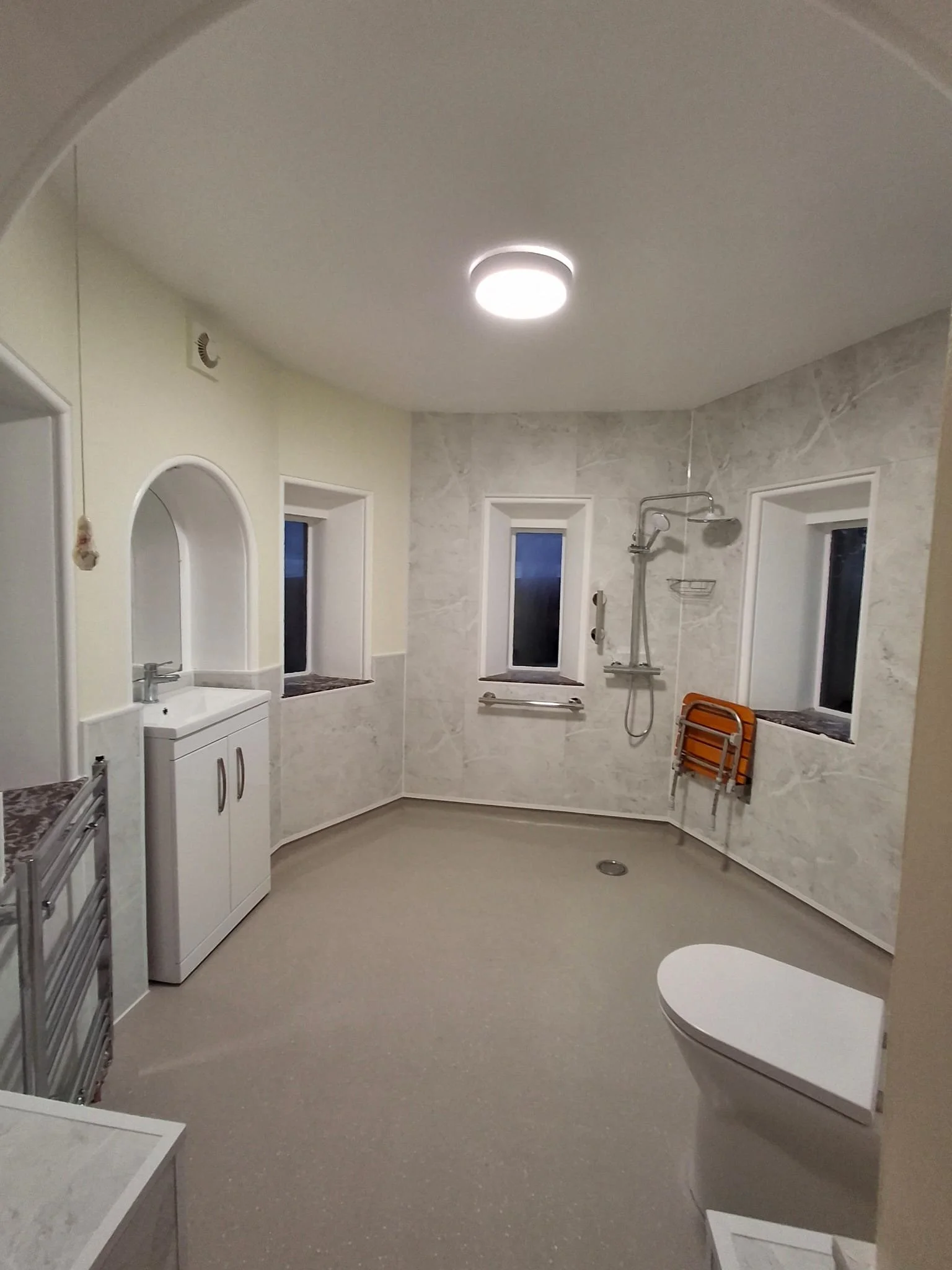 Empty accessible bathroom with a walk-in shower, two small windows, a toilet, and a sink with a cabinet, in a light-colored room with ceiling lights.