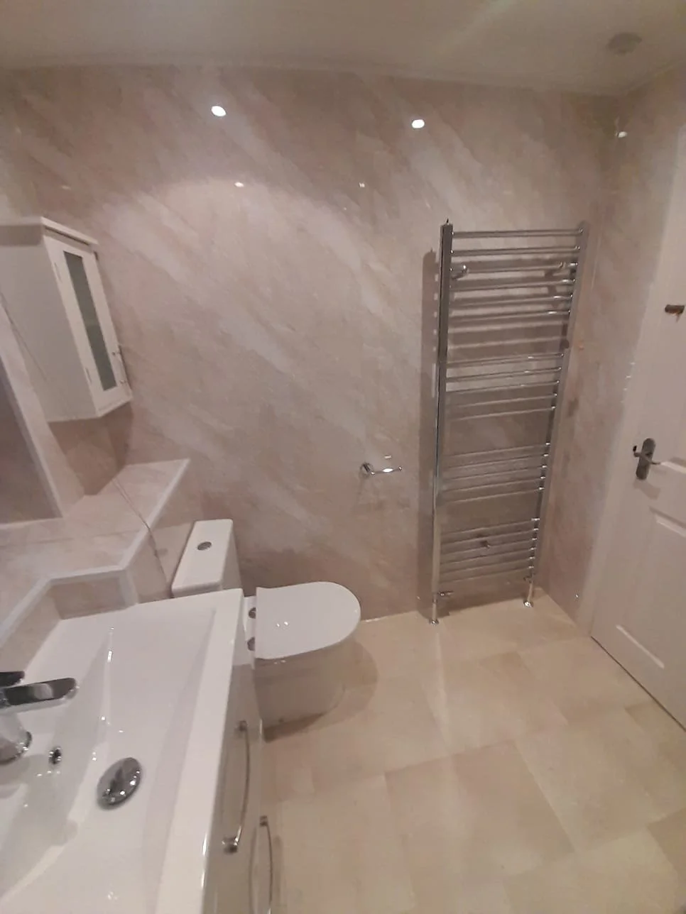 Bathroom with beige tiled floor and walls, white toilet, white sink with silver faucet, wall-mounted white cabinet, heated towel rail, and closed door.