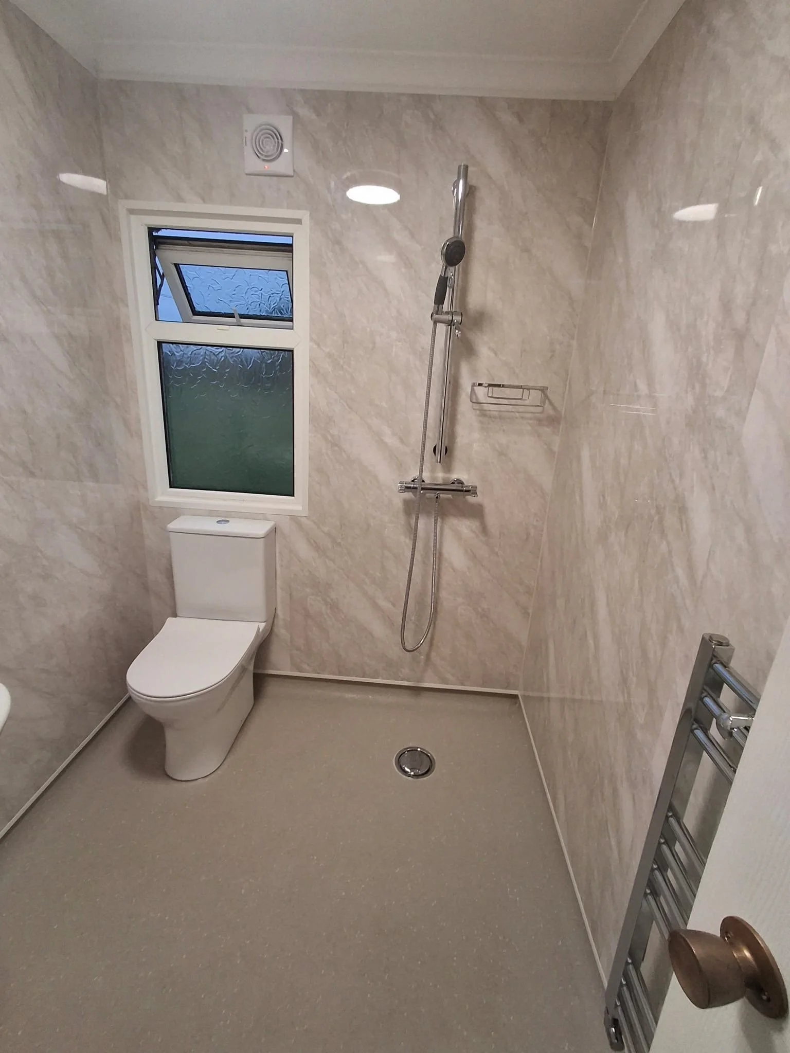 Bathroom with a toilet, a frosted window, a shower with a handheld spray, and a wall-mounted metal rack, all with beige marble walls and a beige floor.